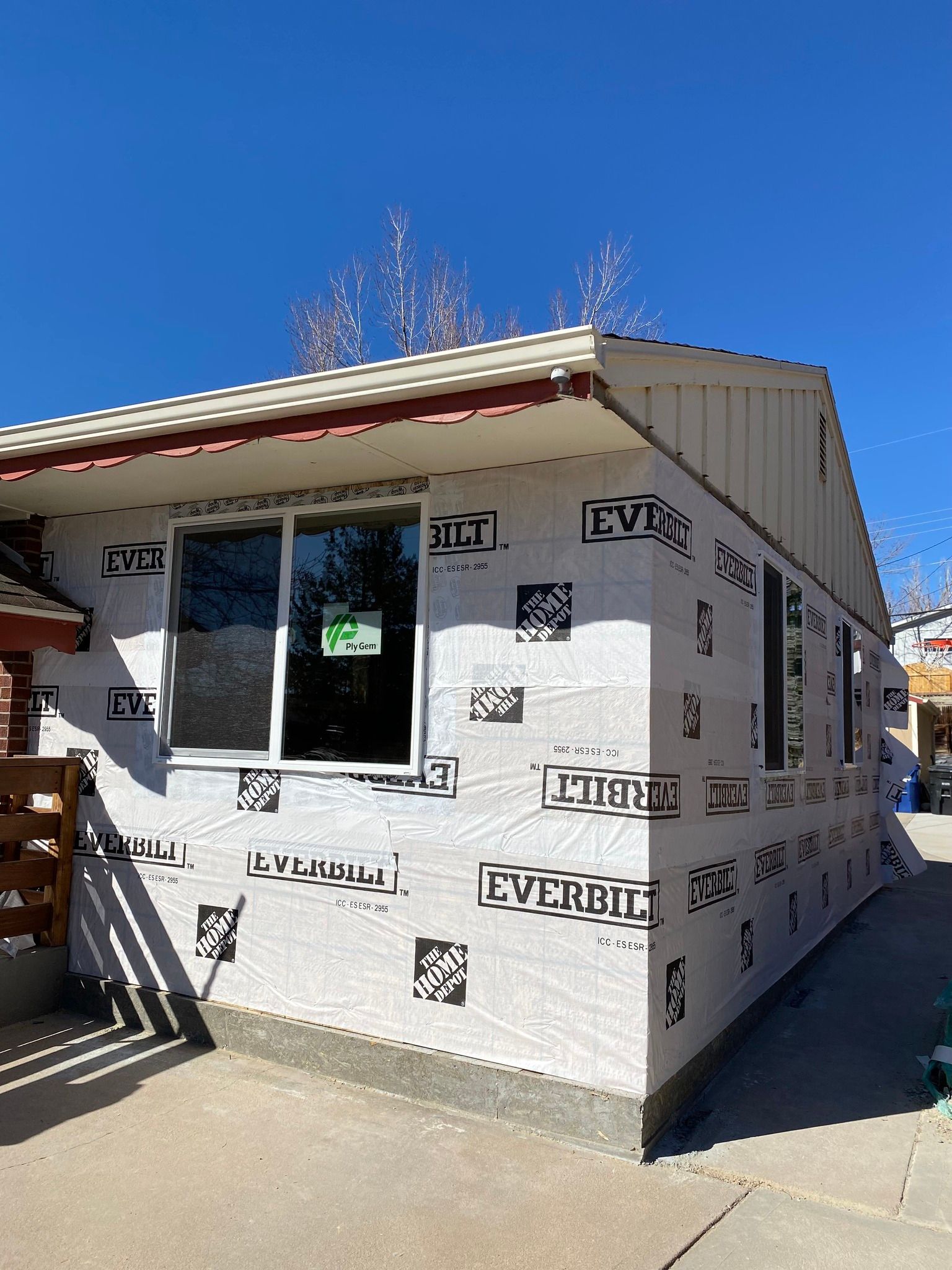 The side of a small house is wrapped in white Everbilt brand house wrap with a visible window under a clear blue sky.