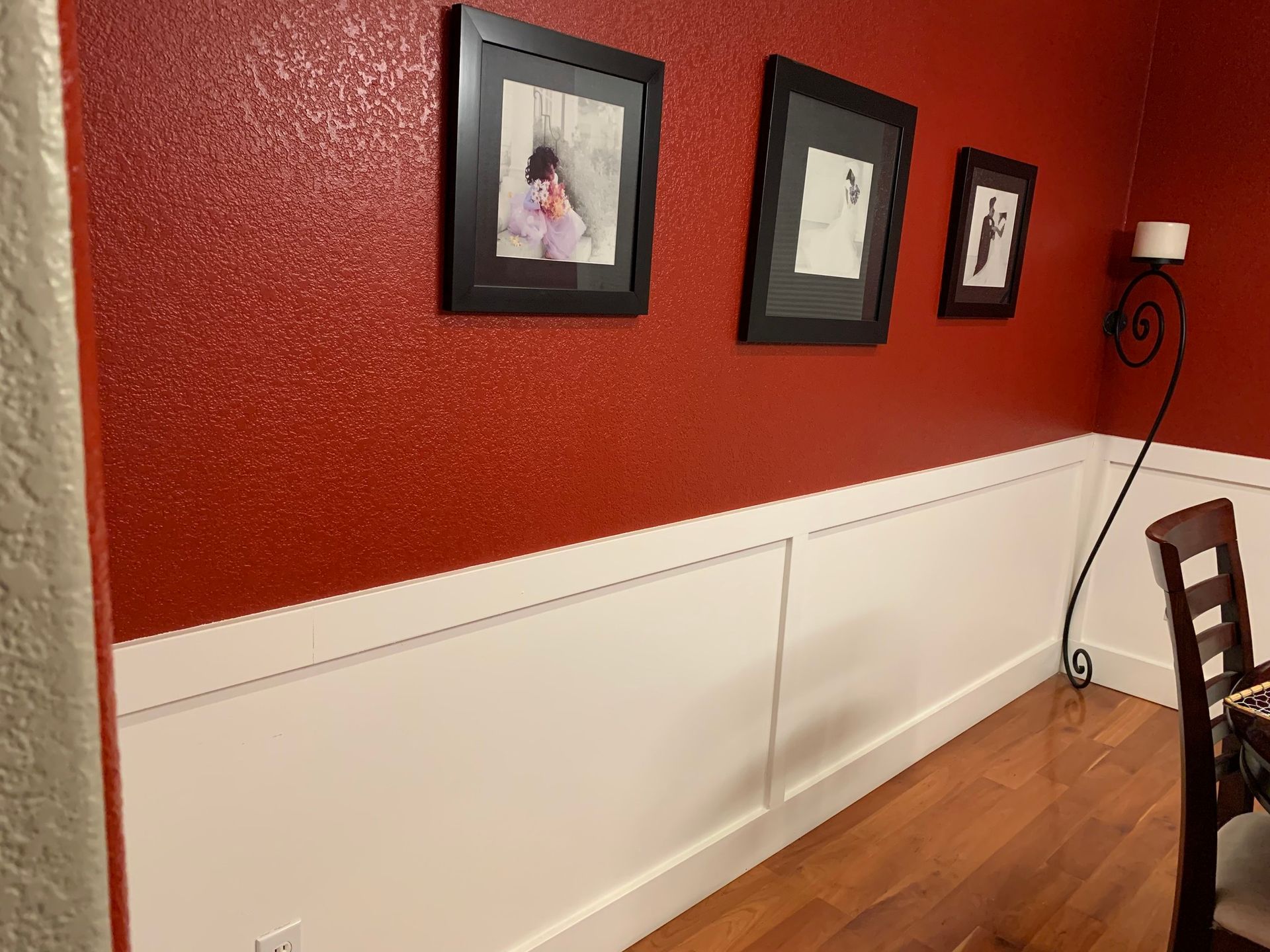 A dining room wall featuring dark red upper walls with three framed pictures above white wainscoting and hardwood floors.