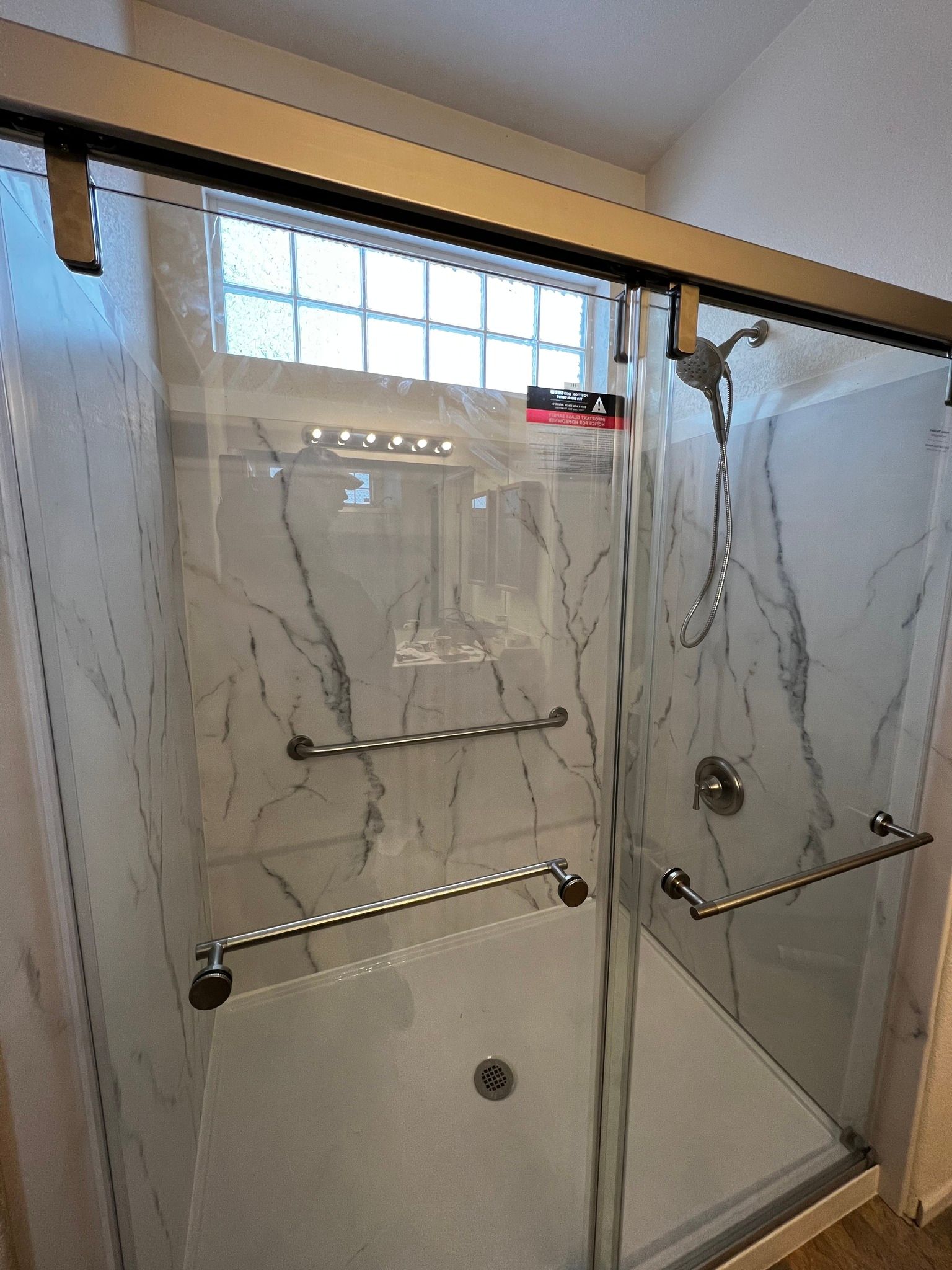 A walk-in shower with marble-patterned walls, a glass sliding door, and multiple metal grab bars installed for support.