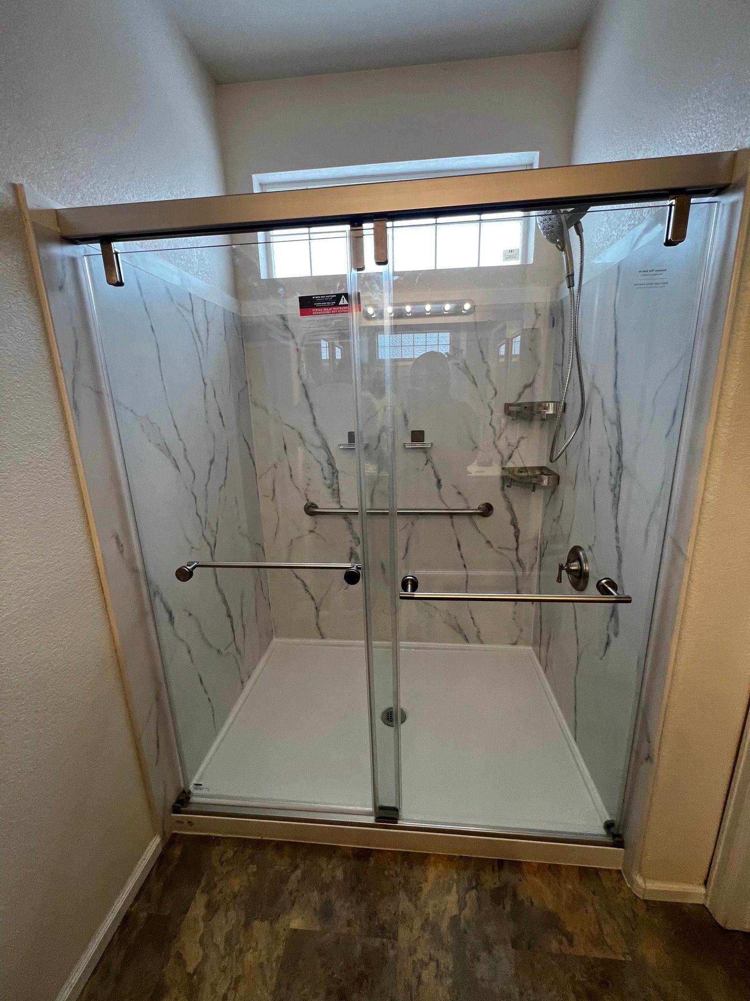 A modern walk-in shower with white marble-patterned walls, a glass sliding door, and two chrome grab bars.