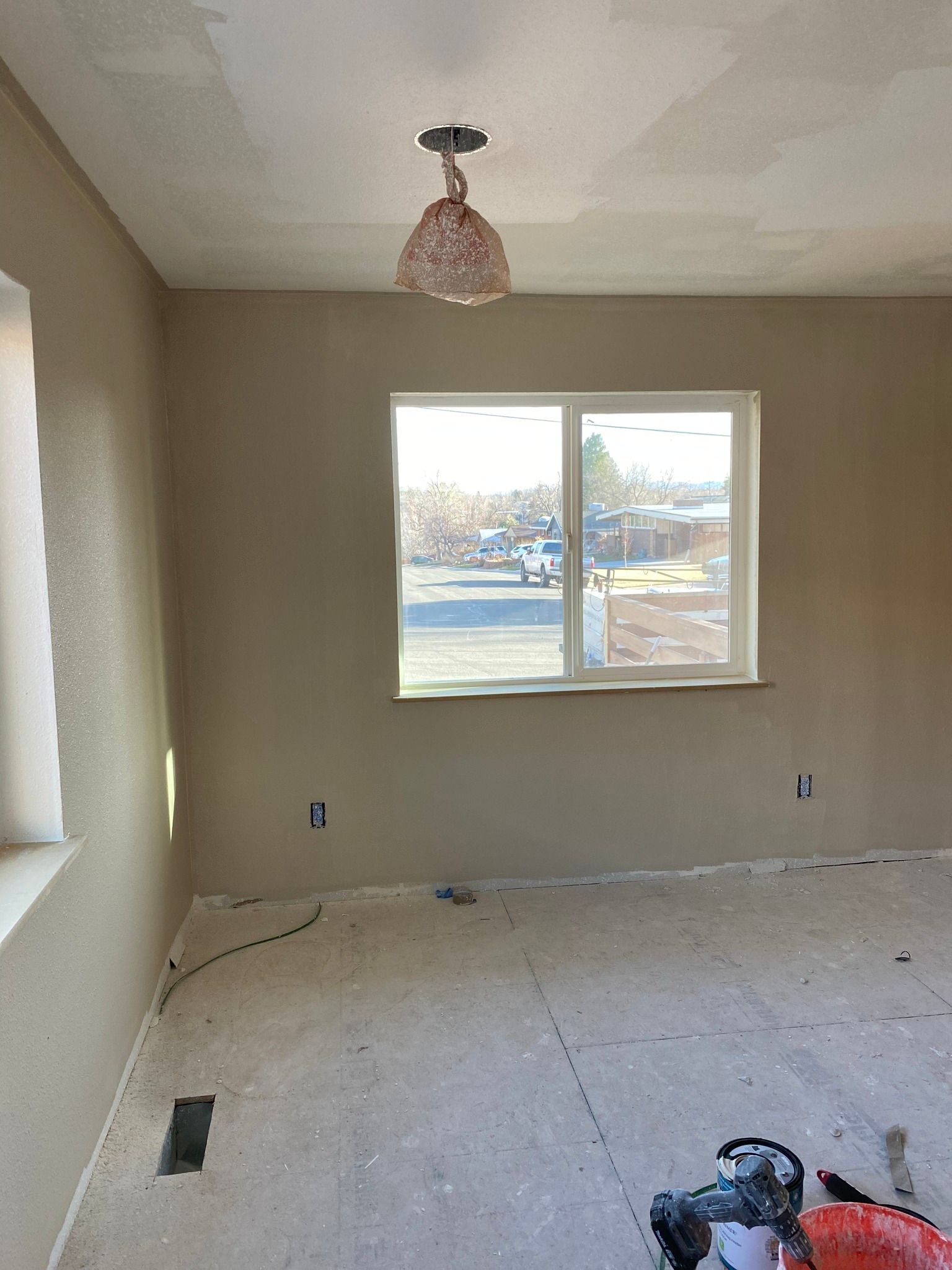 A room under renovation with beige walls, a window, a hanging light fixture, and an unfinished floor with construction gear.