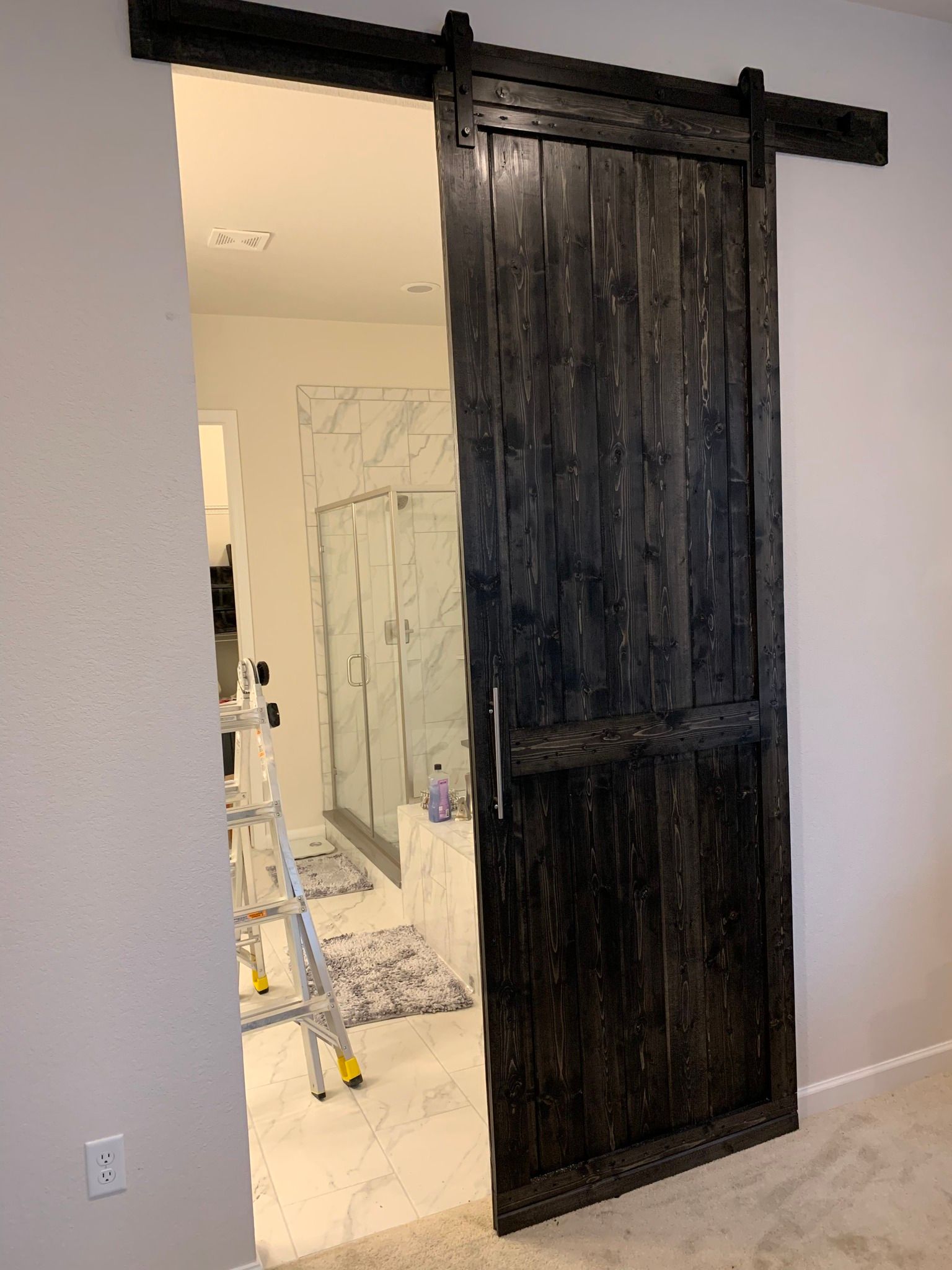 A black, rustic-style barn door hanging on a sliding track in front of a doorway to a bathroom with tile floors.