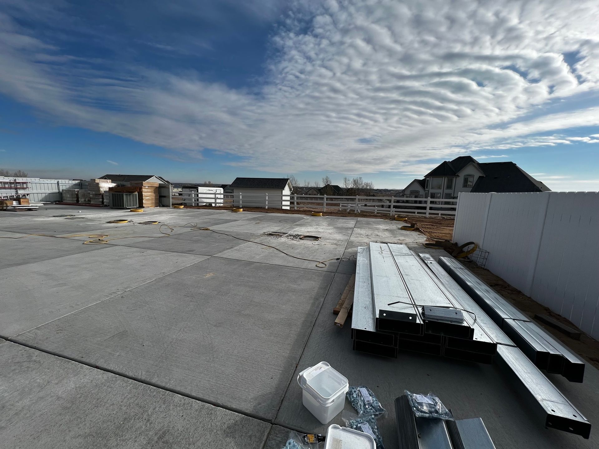 Construction materials stacked on a concrete patio in a backyard under a blue sky with streaky clouds.