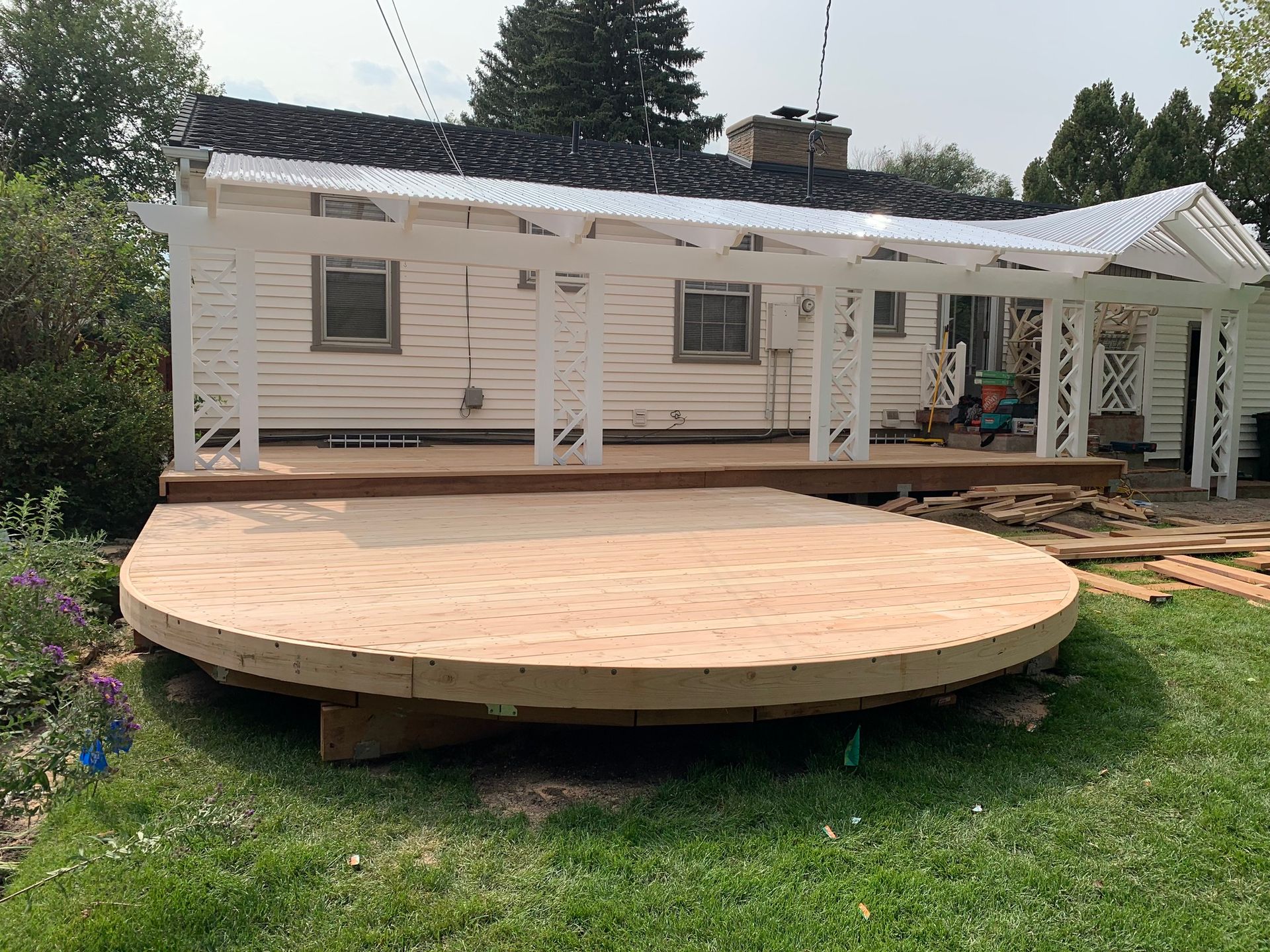A house with white siding features a newly constructed wooden deck with a curved front and an attached white pergola.