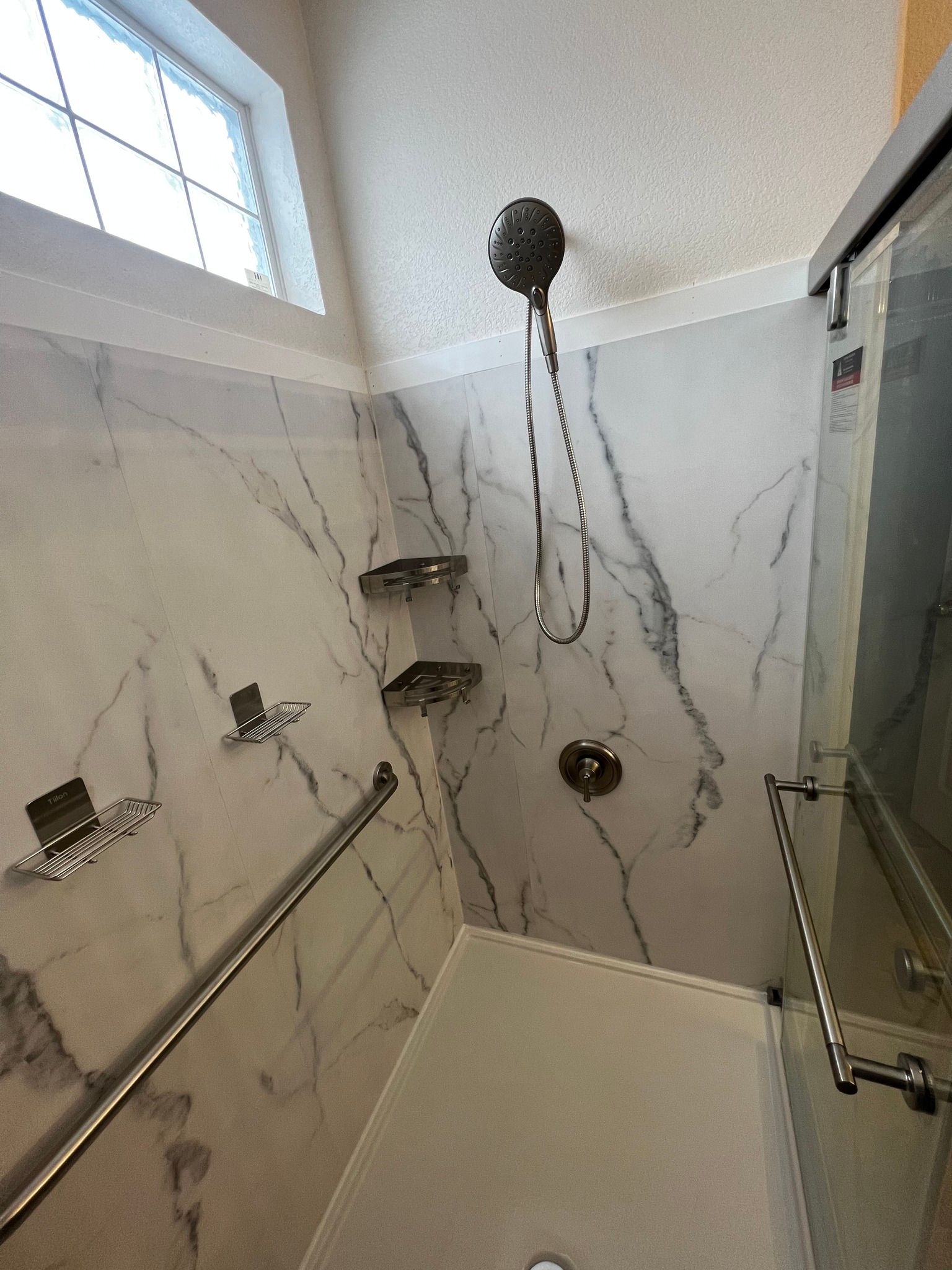 A walk-in shower with white marble-patterned walls, a handheld showerhead, two corner shelves, and a metal grab bar.
