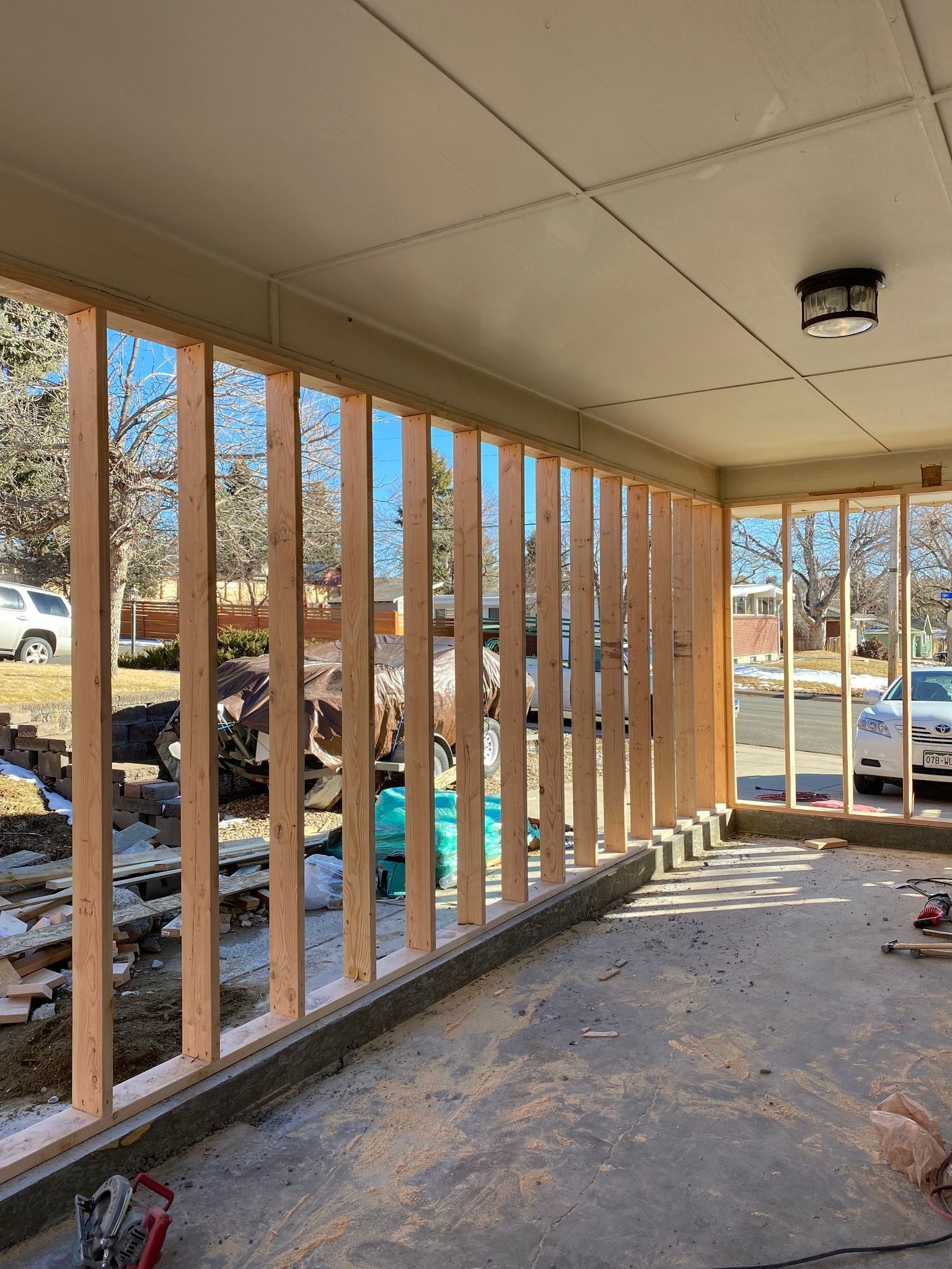 A wooden wall frame under construction on a porch, showing vertical studs installed on a concrete base.