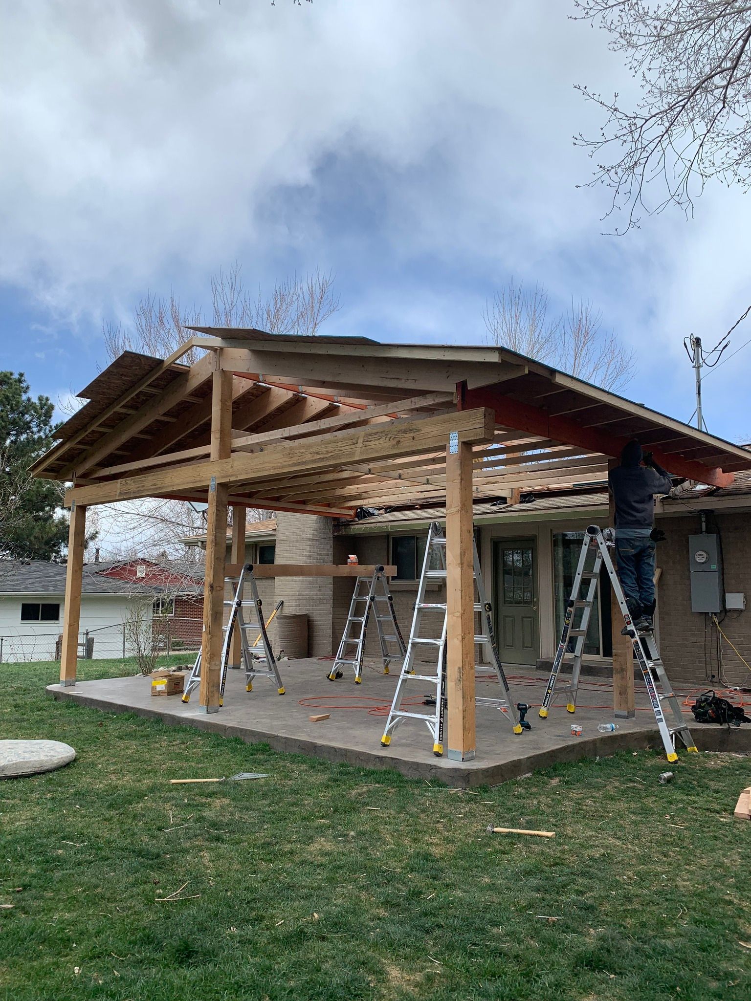A worker stands on a ladder under the wooden frame of a partially built patio cover attached to a brick house.