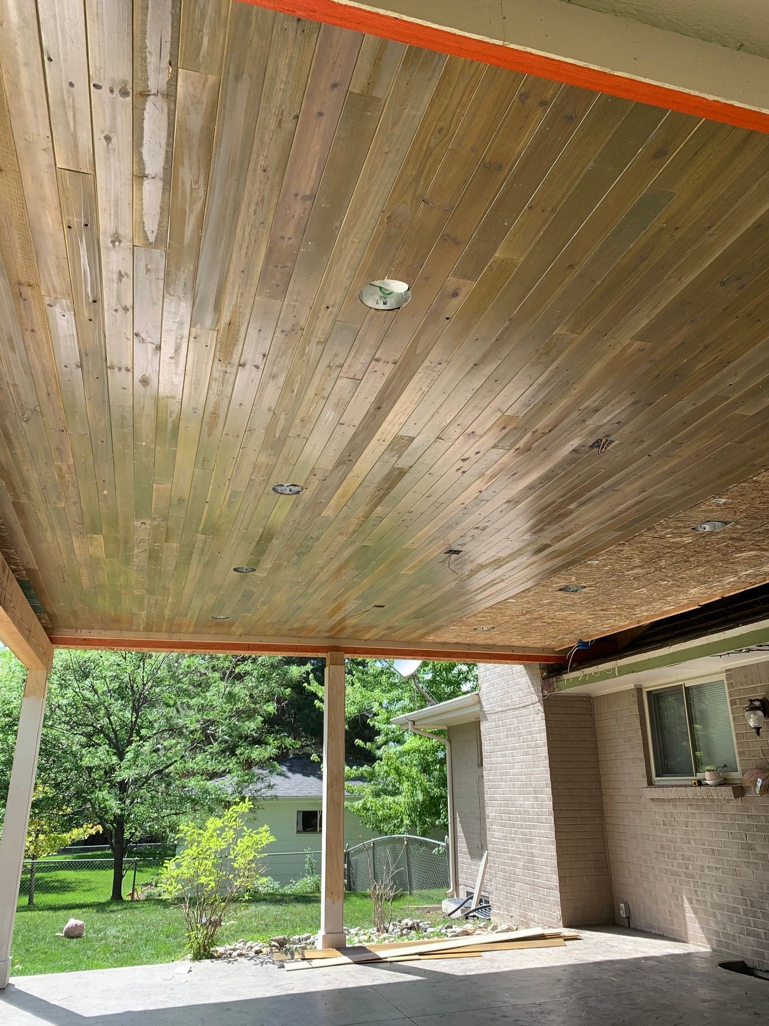 A wooden tongue-and-groove ceiling on a covered patio, showing recessed lighting, support beams, and an outdoor yard view.