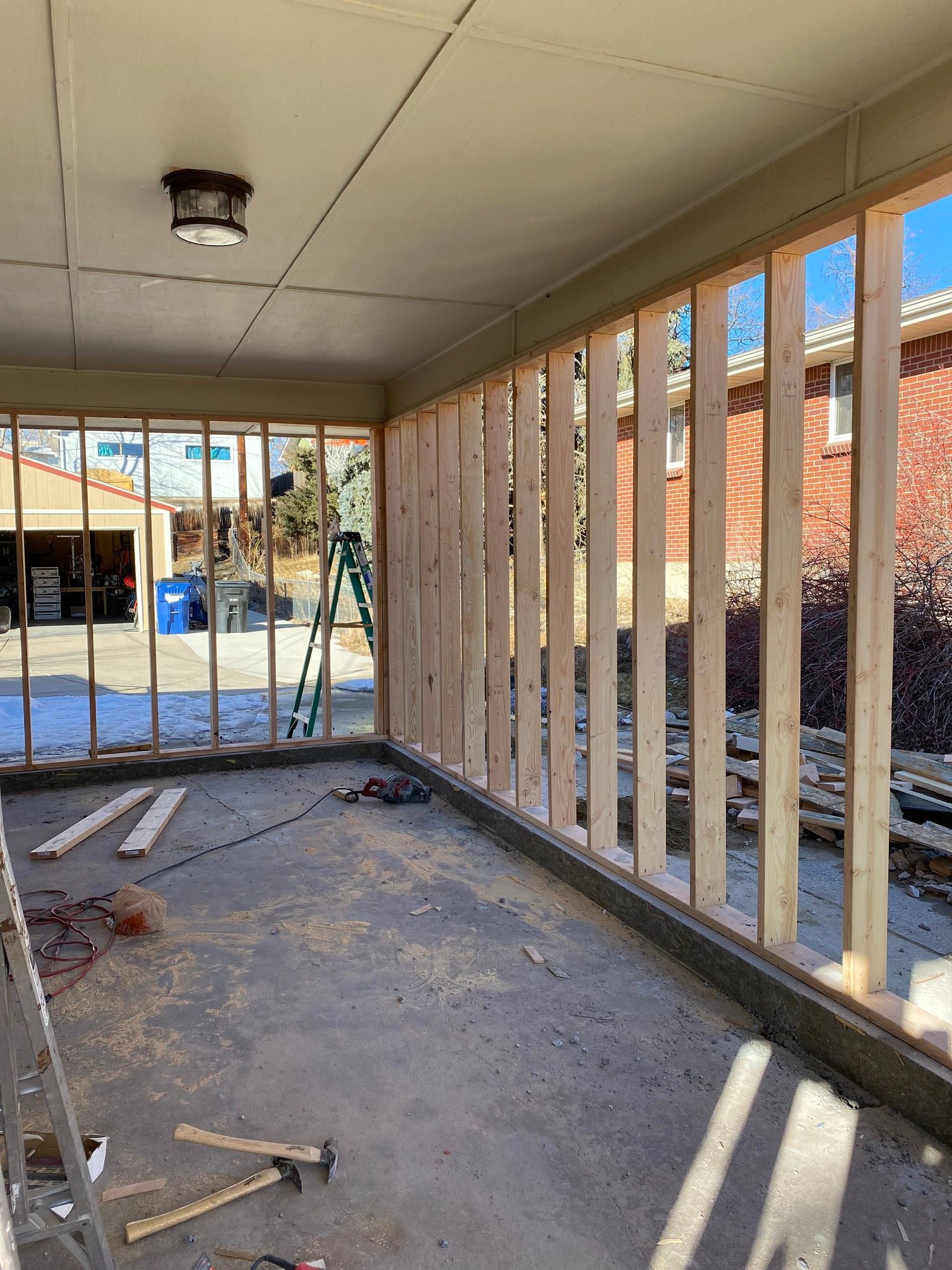 Construction site showing a wooden wall frame being built on a concrete porch under a roof, with a ladder in the distance.