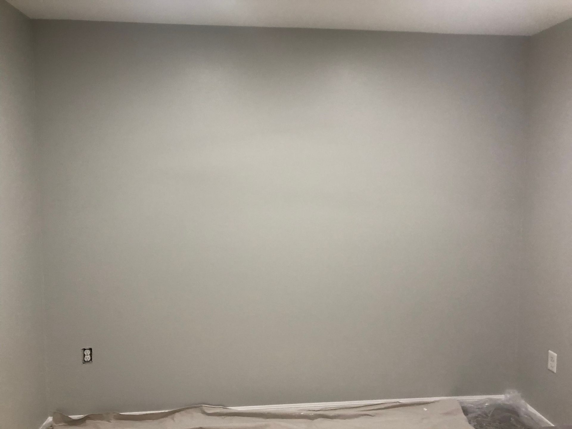 An empty interior room featuring a large, smooth, light-gray wall with two electrical outlets and a floor drop cloth.