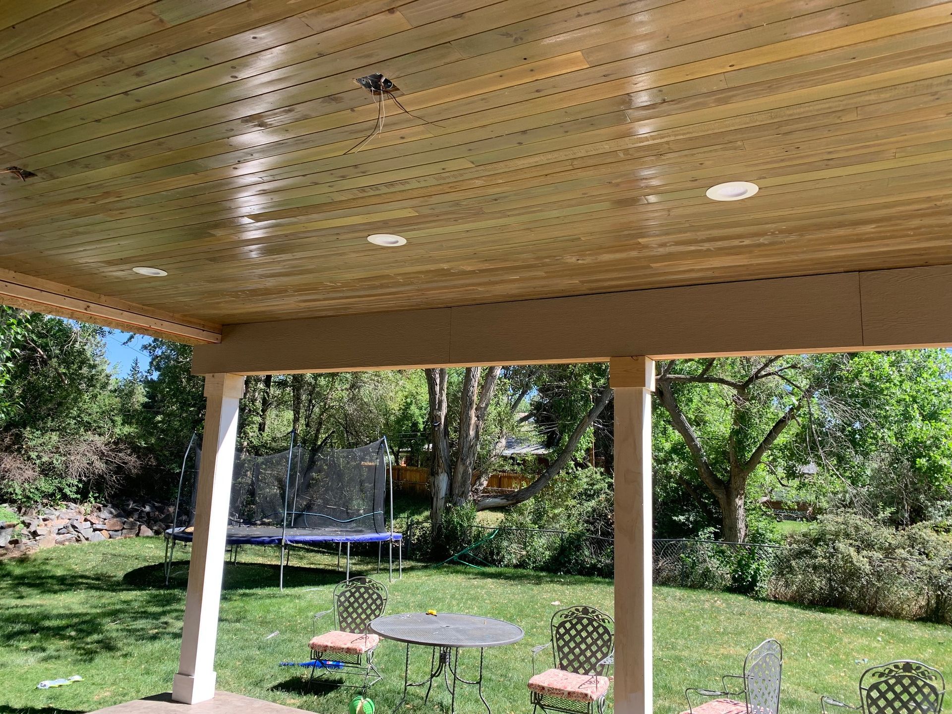 A covered backyard patio with a stained wooden ceiling, recessed lighting, and white posts, overlooking a green lawn.