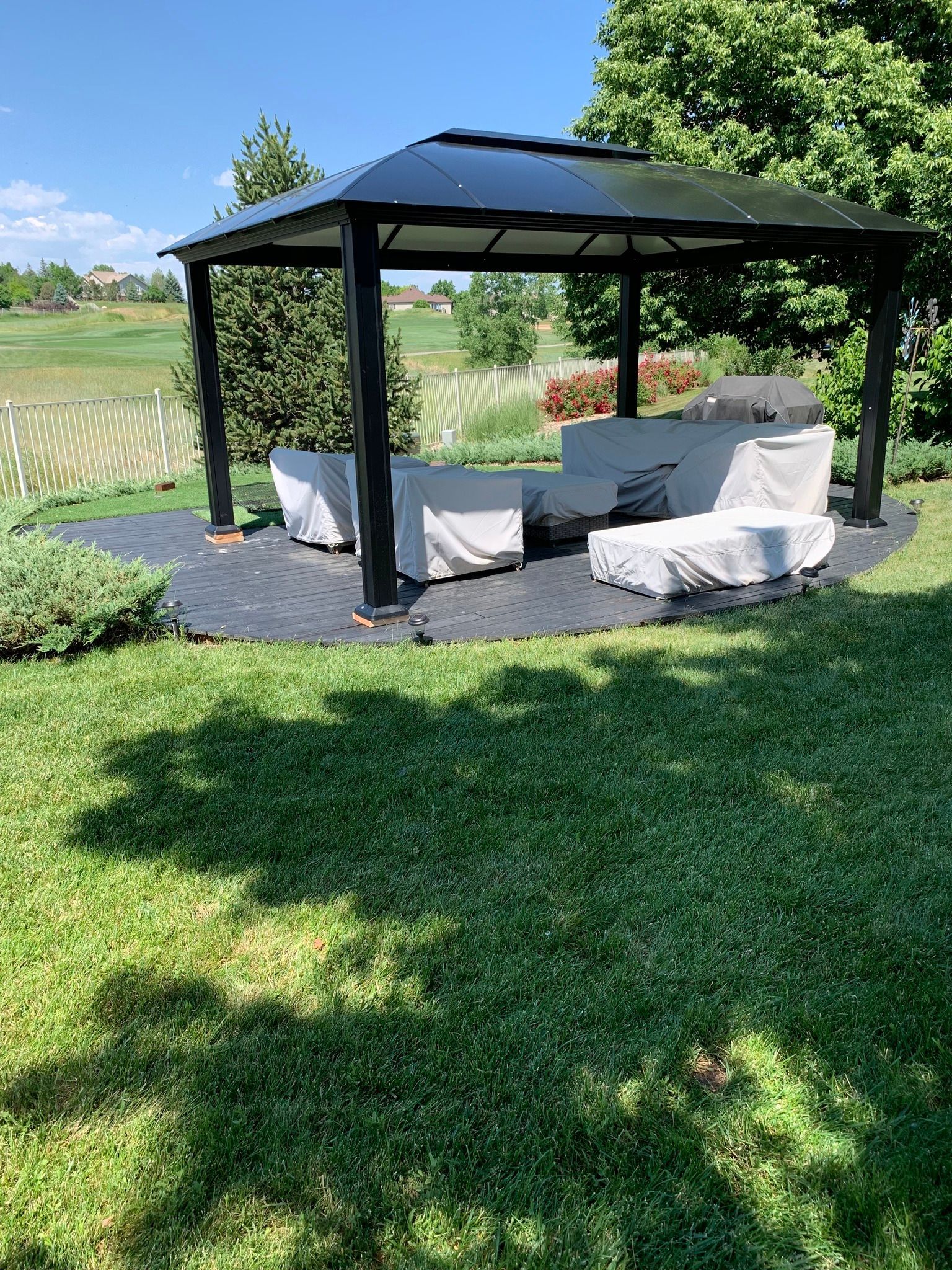 A black metal gazebo sits on a dark stone patio in a grassy backyard, covering patio furniture protected by light covers.