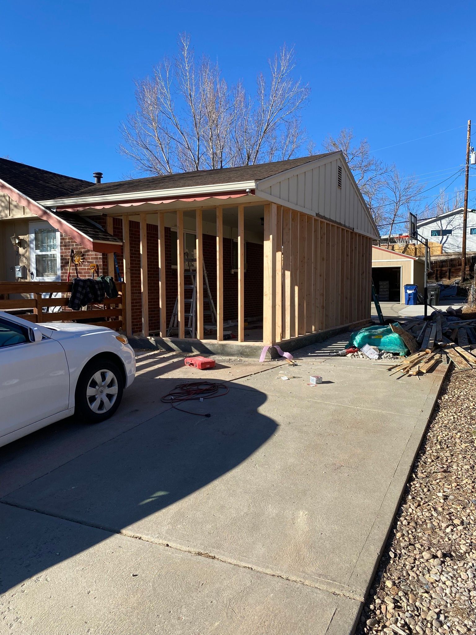 A partially constructed wooden garage frame under a new roof, located next to a house with a white car in the driveway.