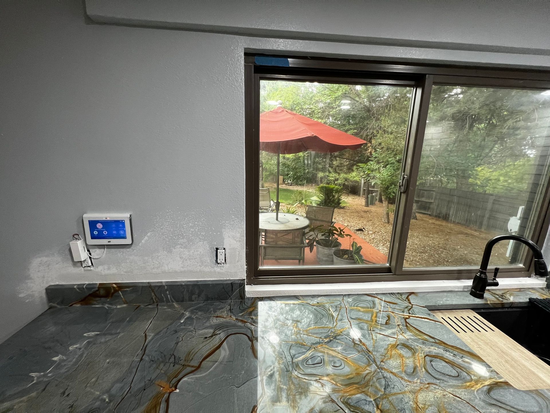 A kitchen counter with dark, veined marble, a smart home display on the wall, and a window view of a patio with an umbrella.