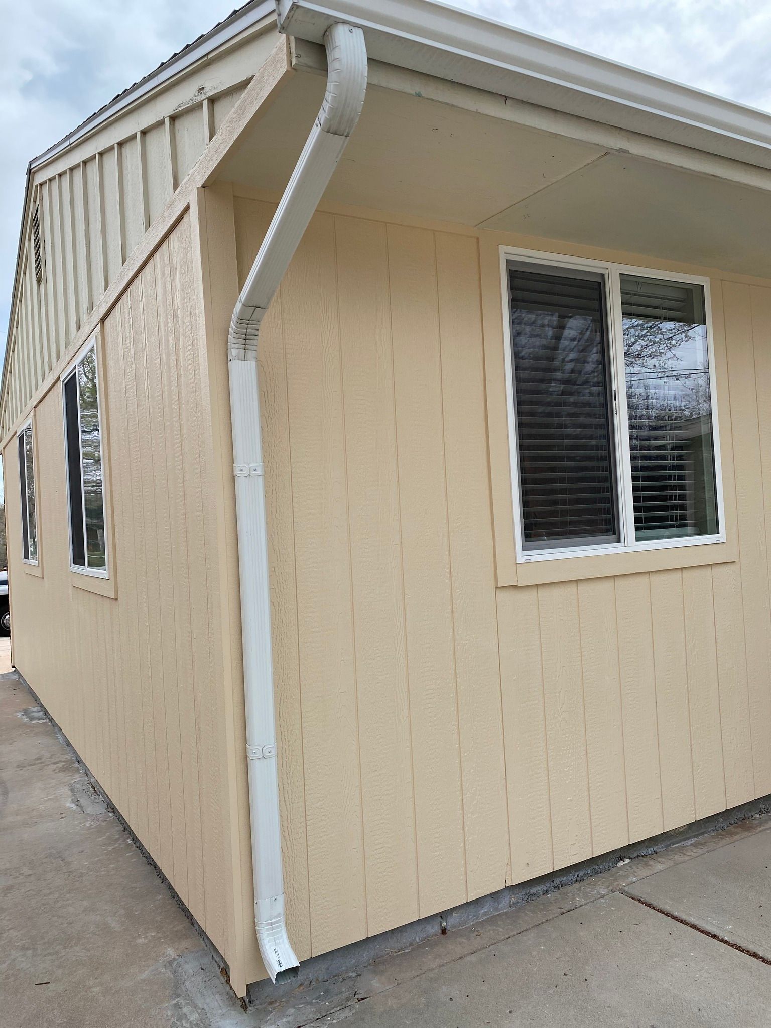 A low-angle view of the corner of a light-yellow house exterior with vertical siding, white trim, and a white downspout.