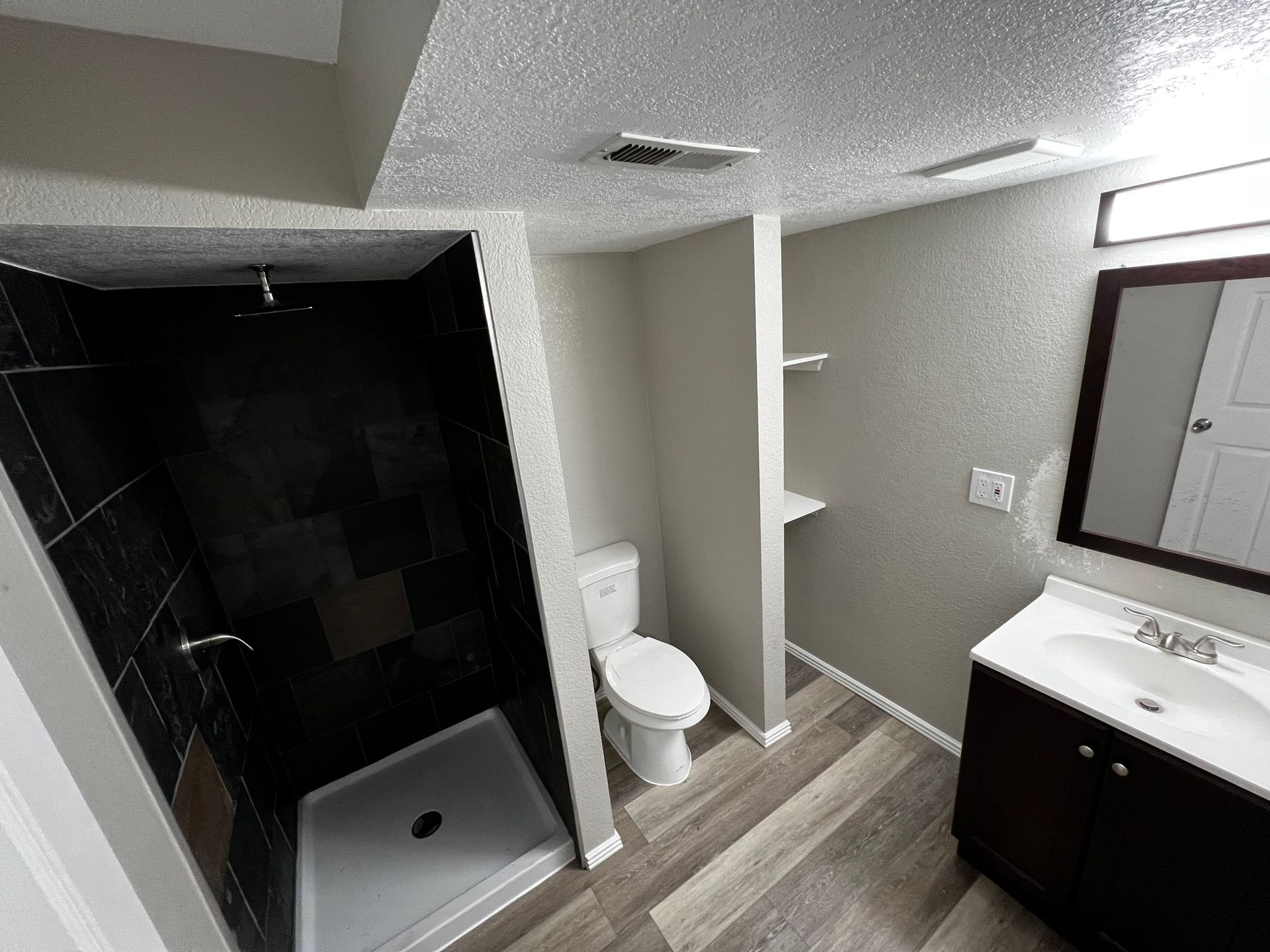 A modern bathroom featuring a black-tiled shower, white toilet in a niche, wood-look flooring, and a dark vanity with mirror.