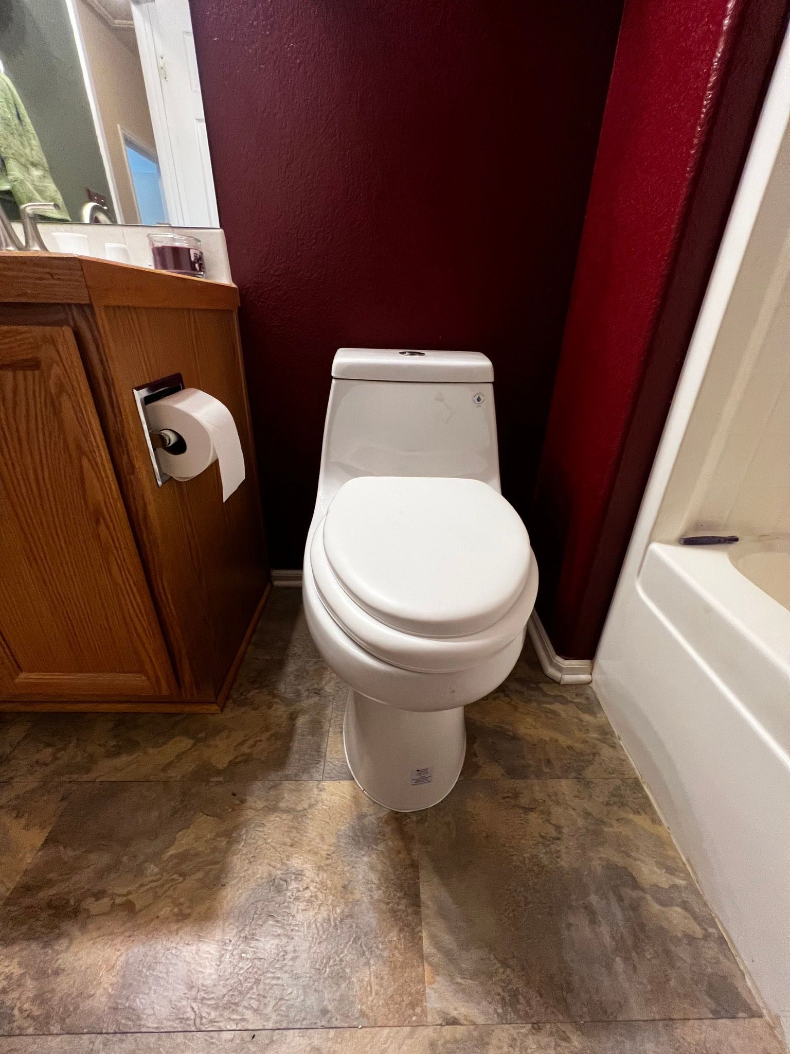 A white toilet sits on a mottled tile floor between a wooden vanity and a shower wall against a deep red wall.