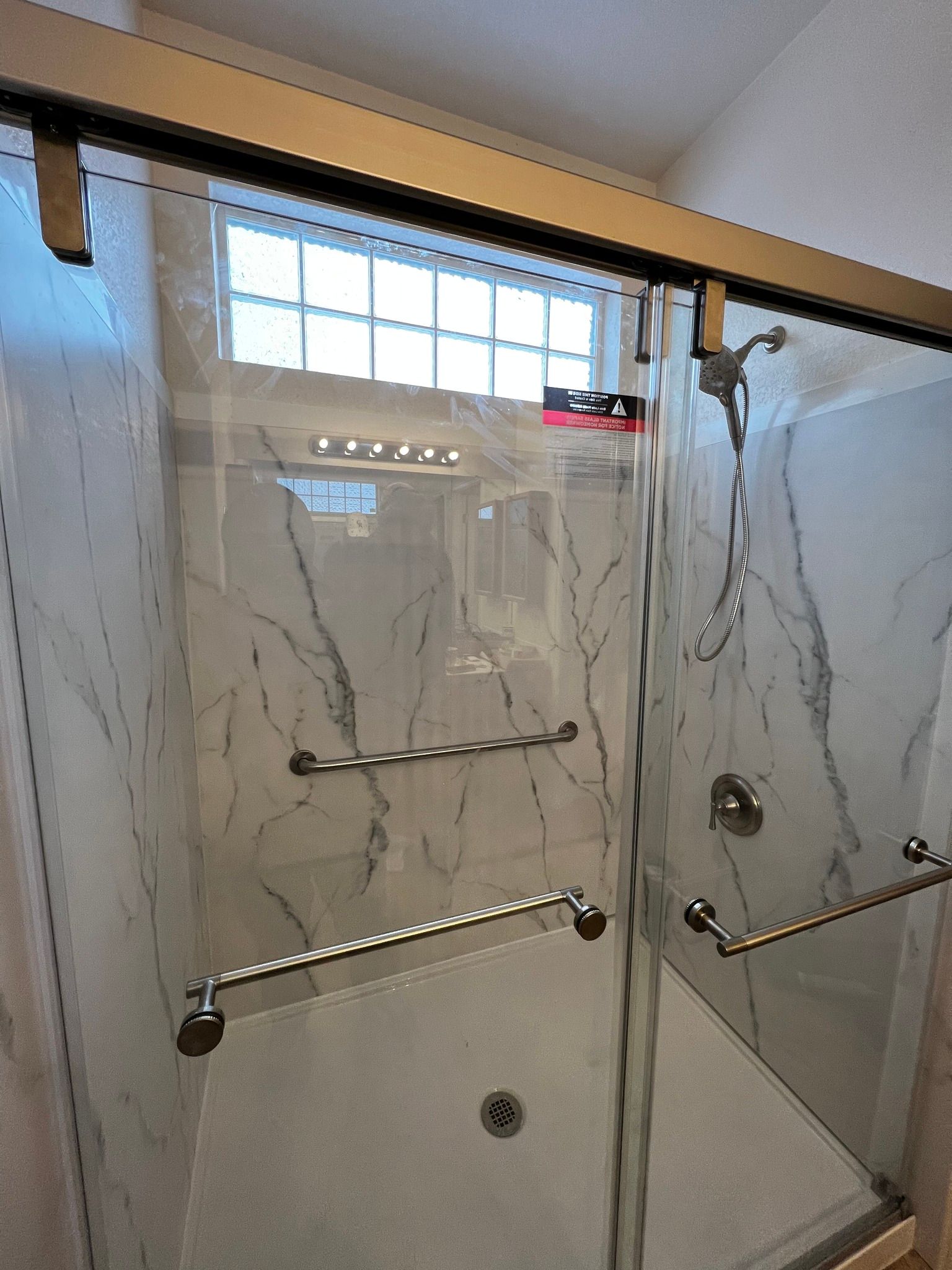 A walk-in shower with marble-patterned walls, a glass sliding door, and two chrome safety grab bars installed inside.