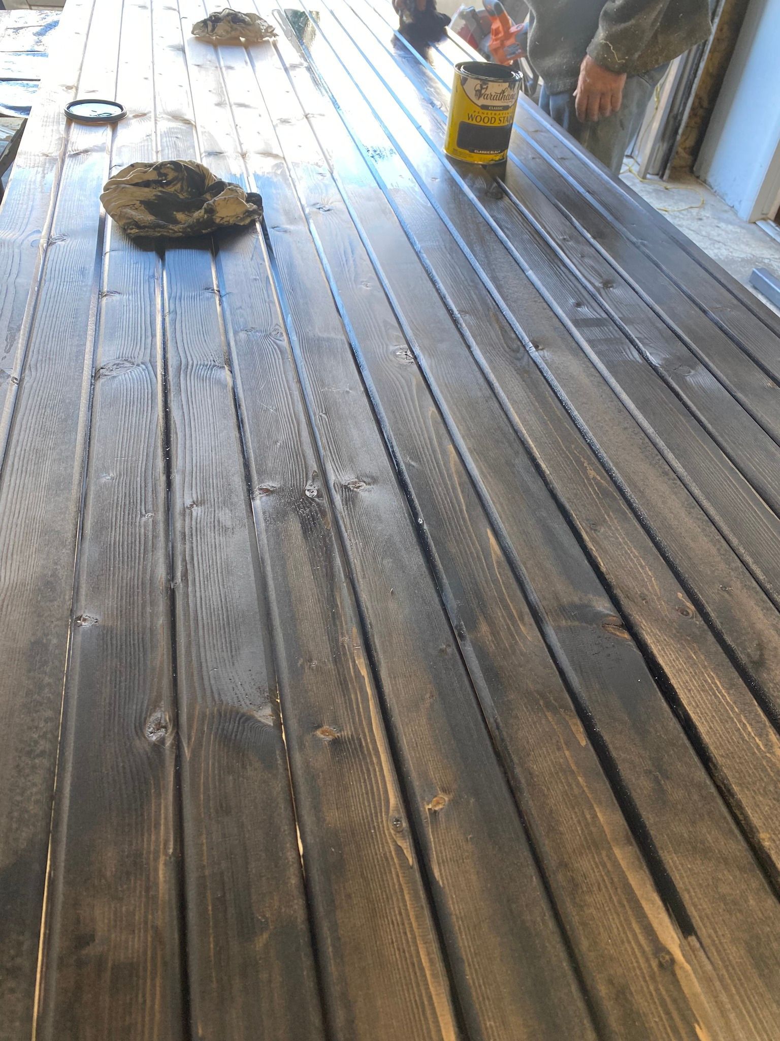 A person applies dark stain to a wooden deck, with a paint can nearby on the planks.