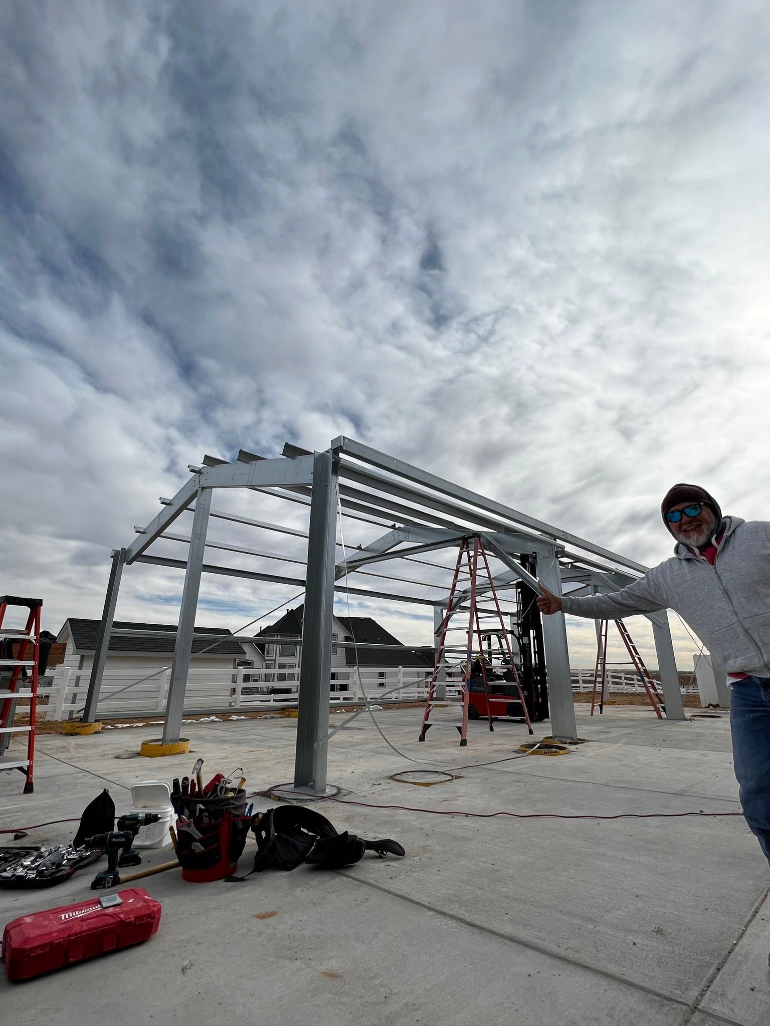 A person stands in the foreground as construction workers assemble the metal frame of a building under a cloudy sky.