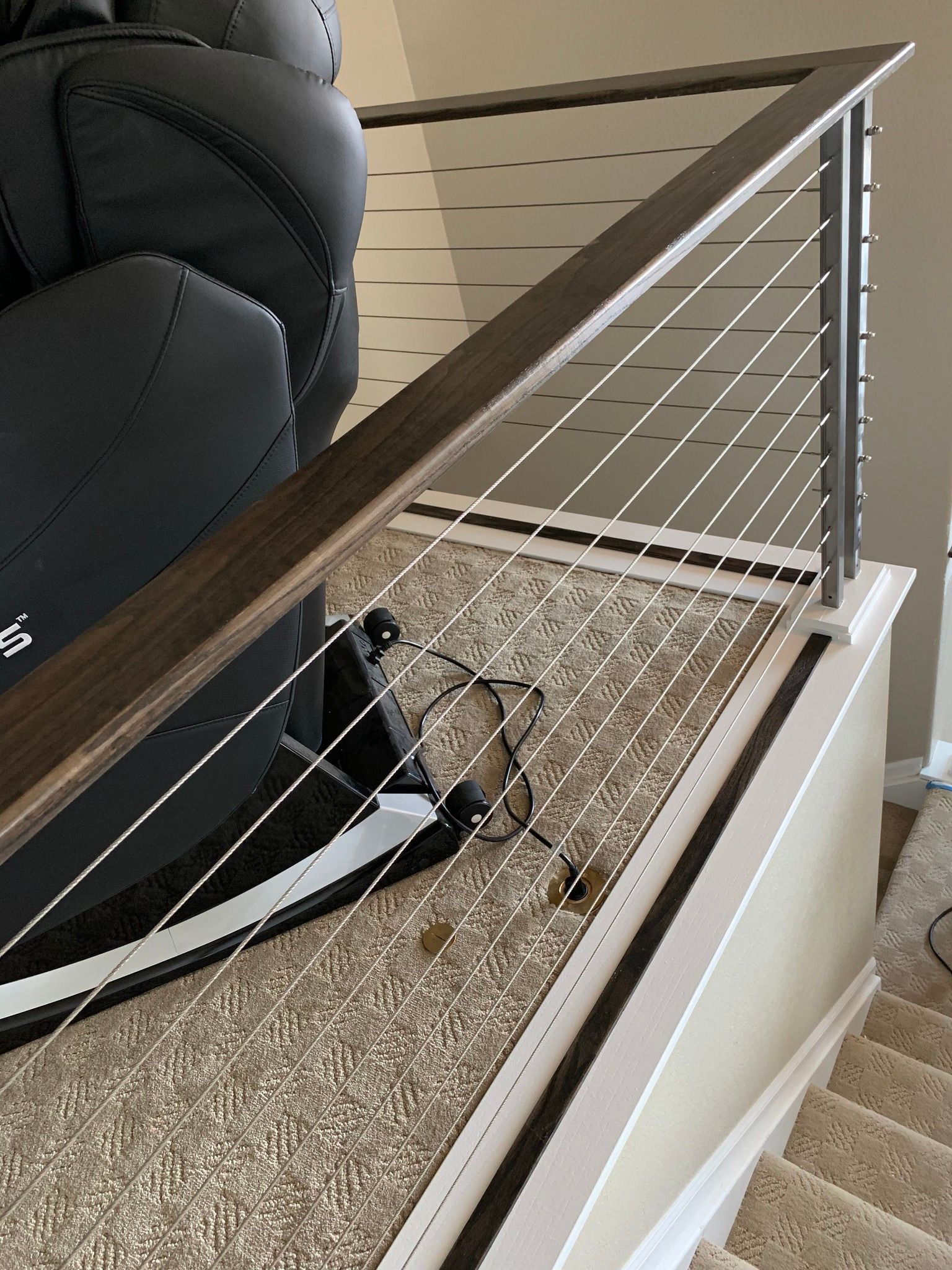 A dark recliner sits near a modern staircase with a wooden handrail and thin horizontal cable railings.