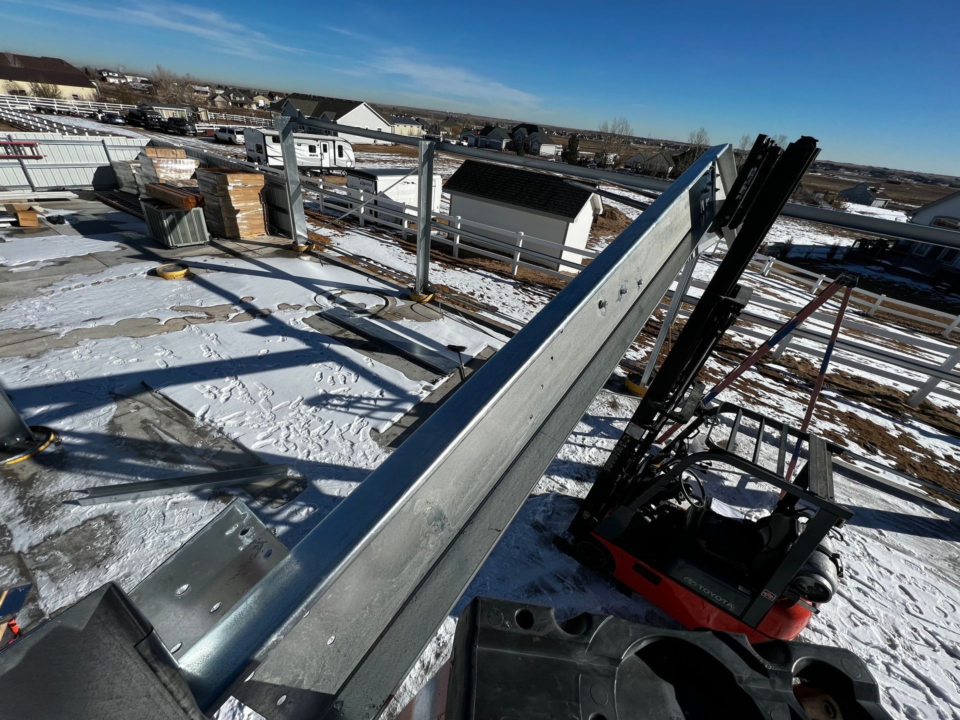 A diagonal view of a large metal structural beam on a snowy construction site with a red forklift in the distance.
