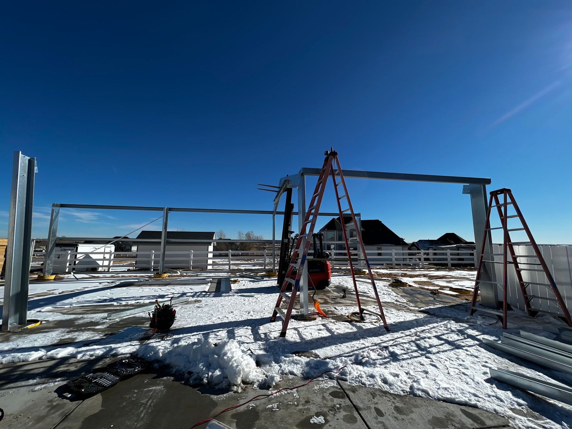 Steel framing for a new structure under construction on a snowy, sunny day, with ladders and machinery nearby.