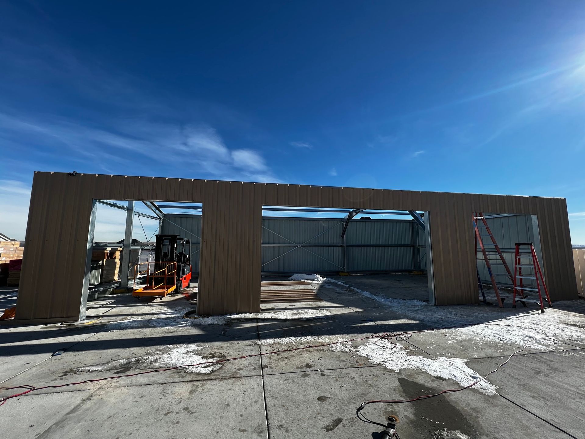 Construction of a brown metal-sided building under a bright blue sky, with a forklift and ladder visible at the entrance.