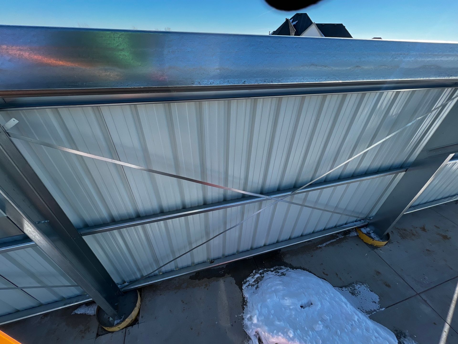 A section of corrugated metal fencing with a diagonal wire cross-brace attached to a metal post on a snowy concrete surface.