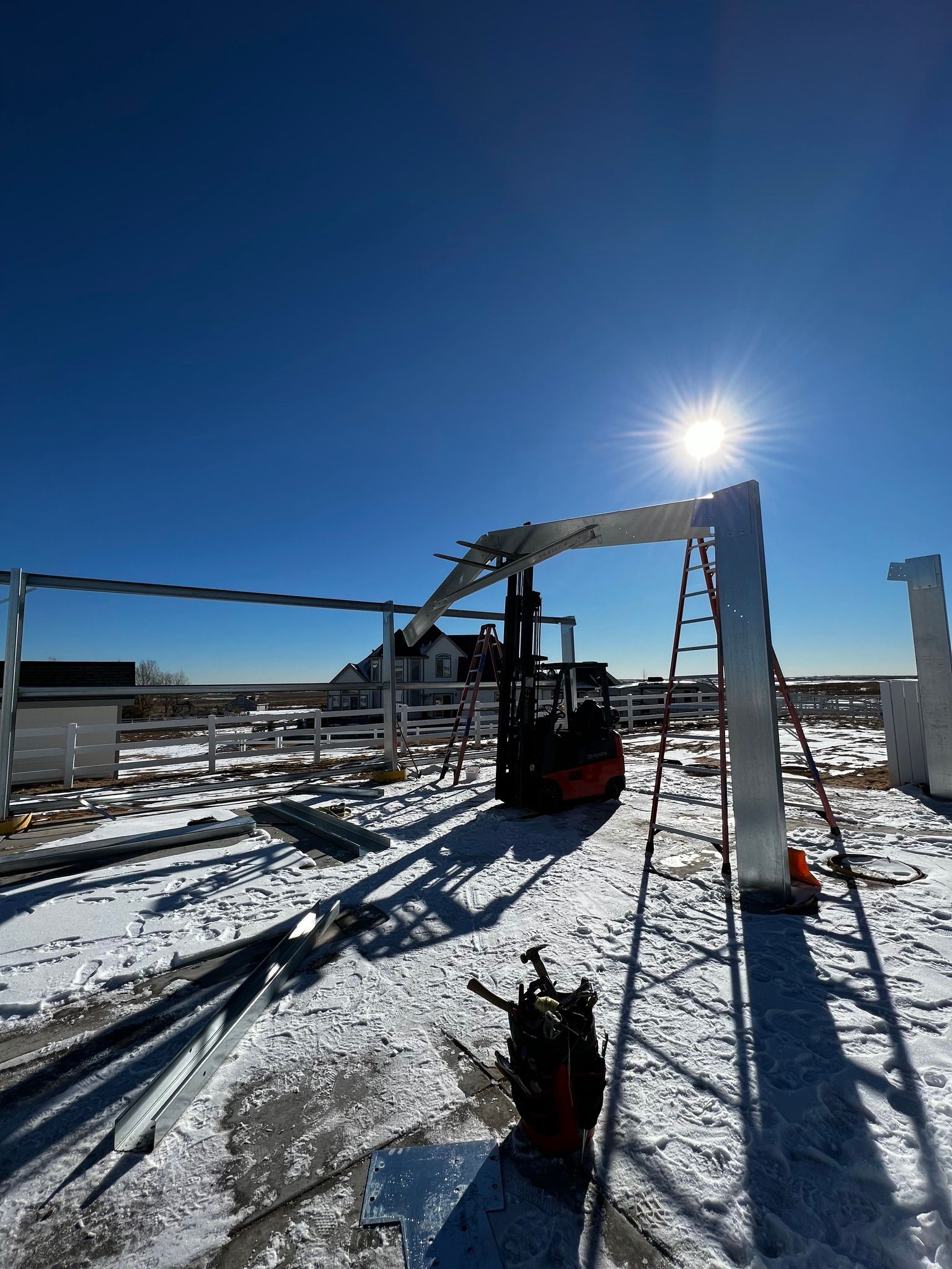 Construction site with a forklift lifting a metal beam under a bright sun, set on a snowy field with a house in distance.