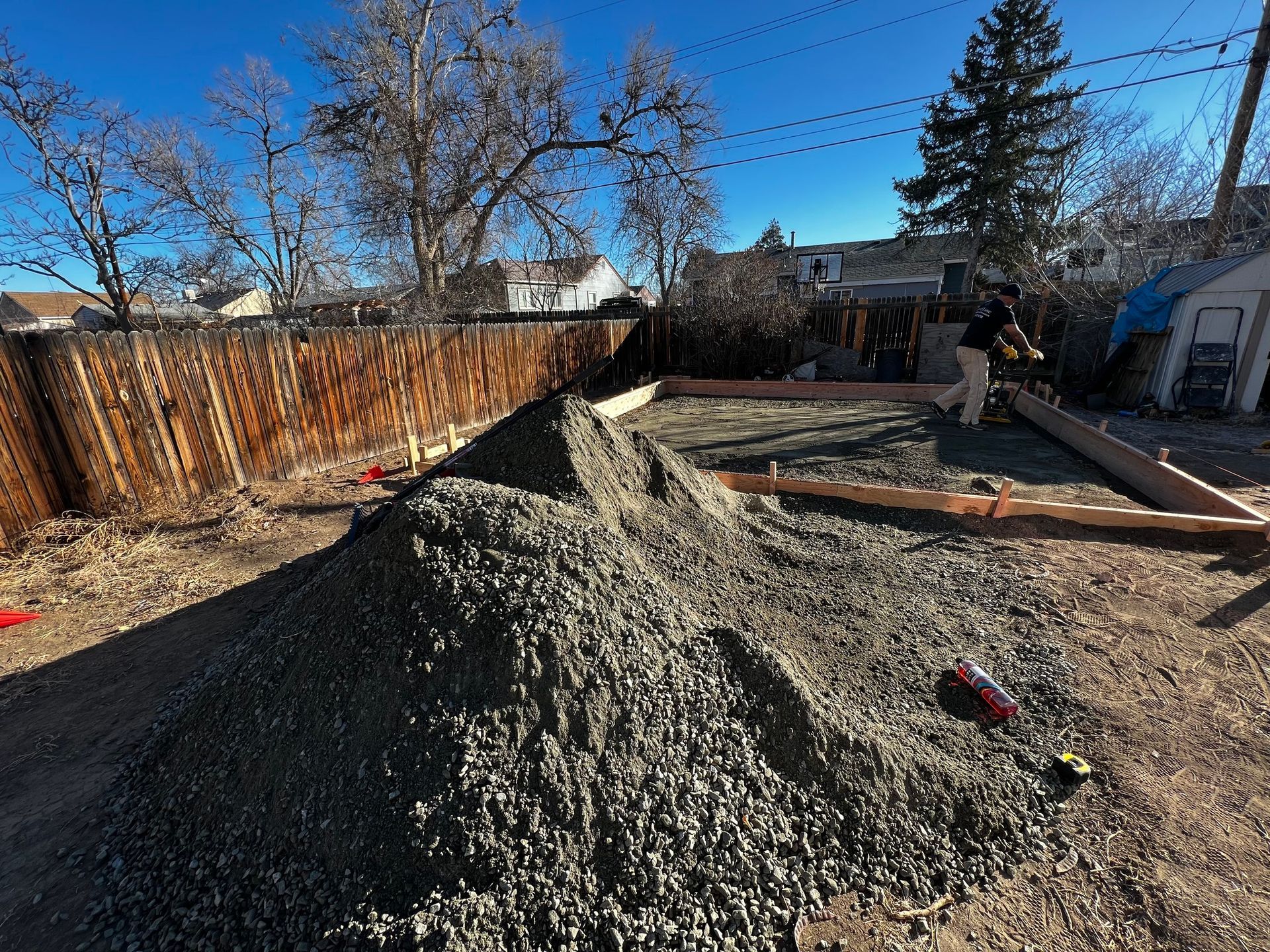 A pile of gravel sits in a backyard next to a wooden rectangular frame under construction on a clear, sunny day.