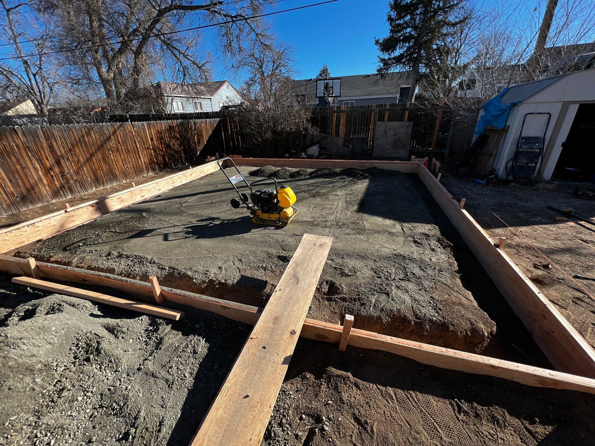 A yellow plate compactor sits inside a wooden form frame filled with packed gravel for a concrete pad project.