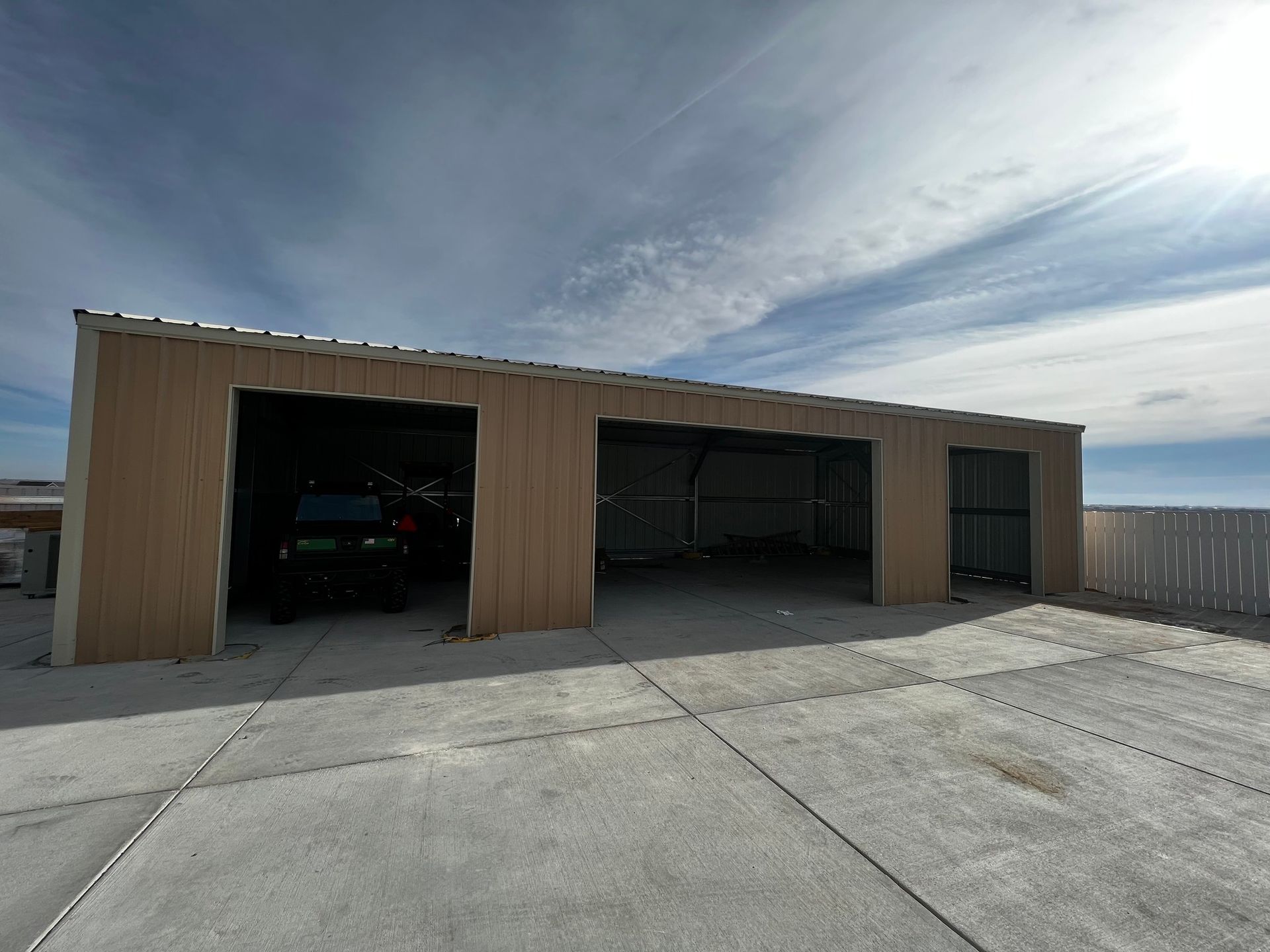 A wide, tan metal storage building with three open bays under a sunny, cloudy sky, situated on a concrete slab.