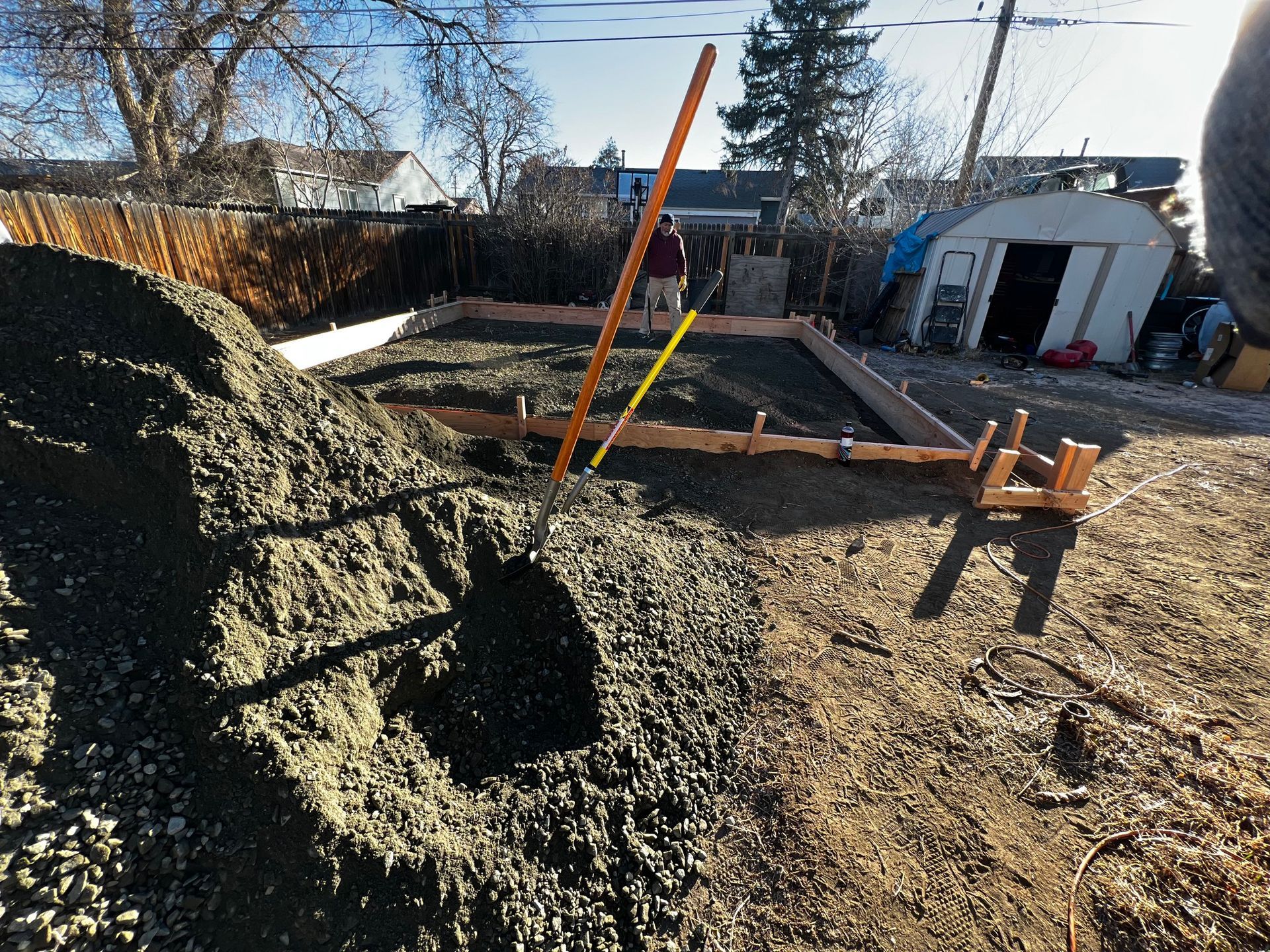 A backyard construction site featuring a wooden rectangular frame filled with gravel, next to a large pile of gravel.