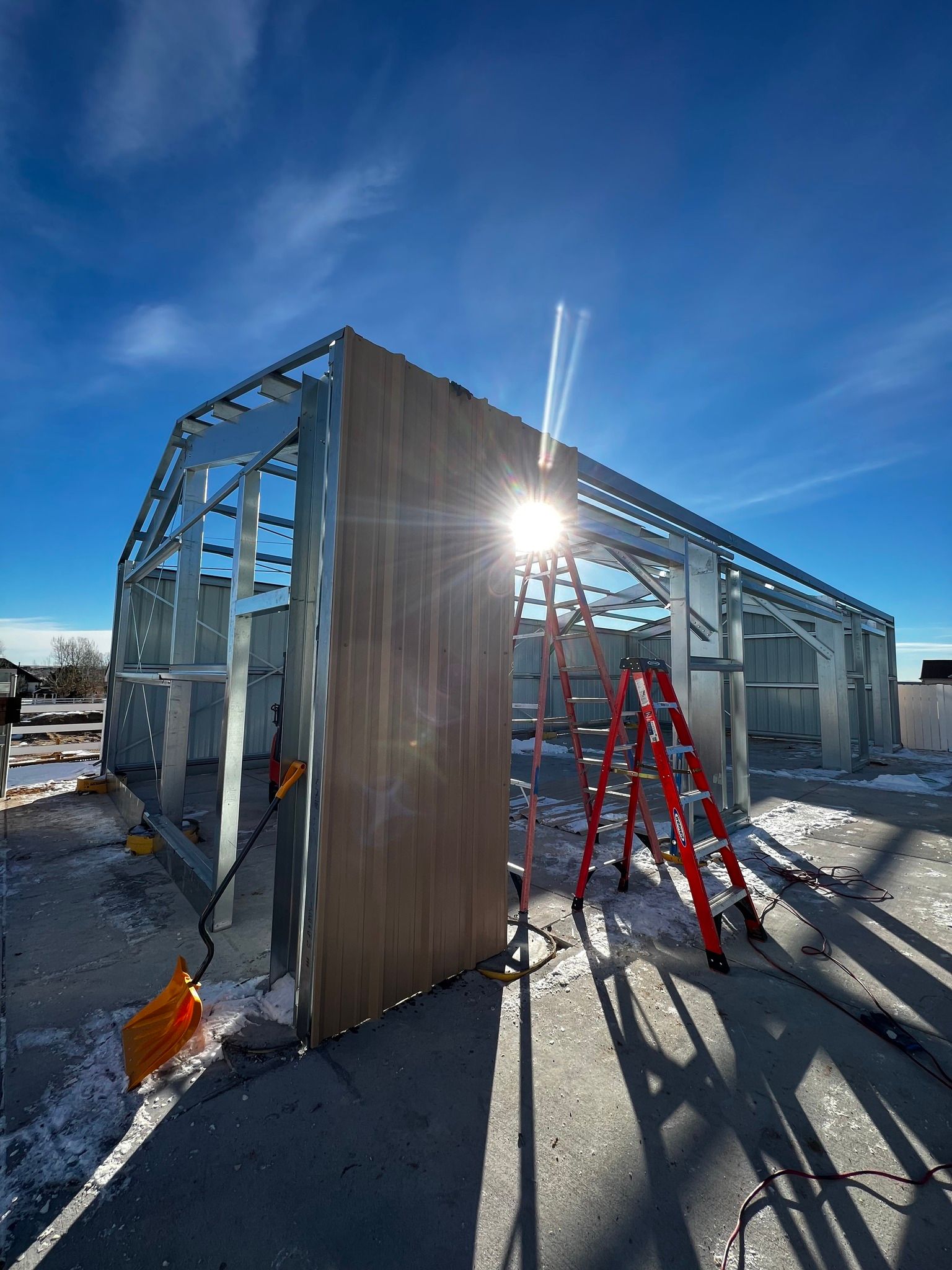 Metal frame of a building under construction on a snowy day, with a red ladder and bright sunburst in the background.