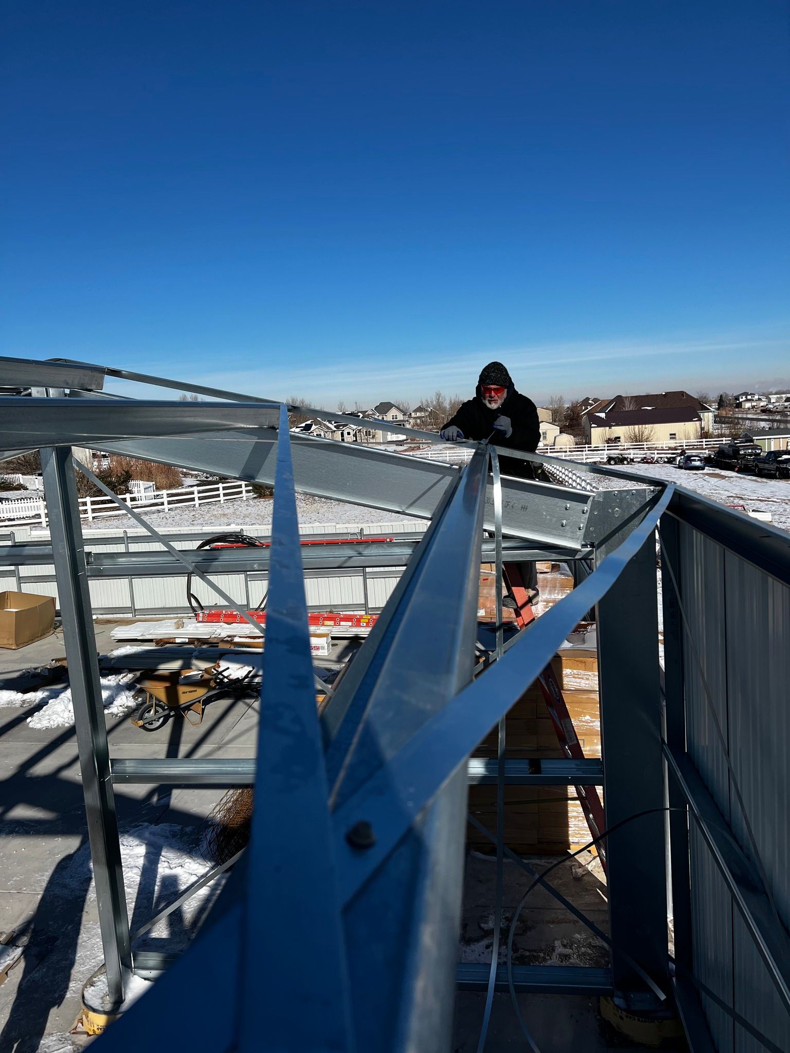 A person in a black hoodie works on assembling a metal building frame outdoors on a sunny, snow-covered day.