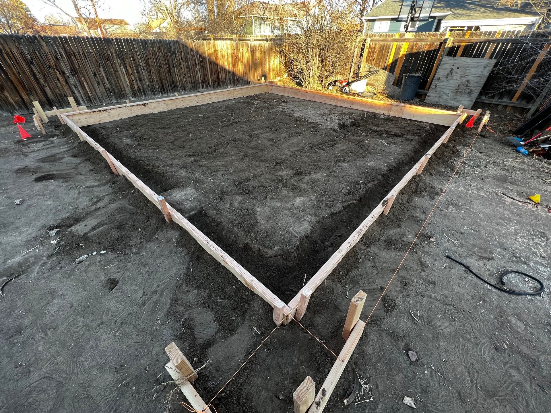 A square wooden concrete form sits in a dirt backyard, prepared for a foundation pour near a wooden fence.