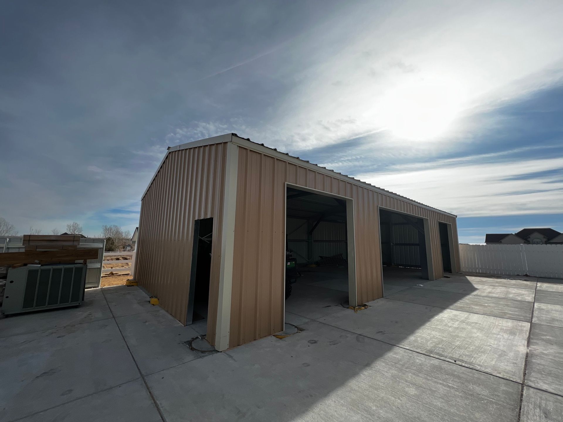A metal garage building with tan siding and three open bays, set on a concrete pad under a sunny, cloudy sky.