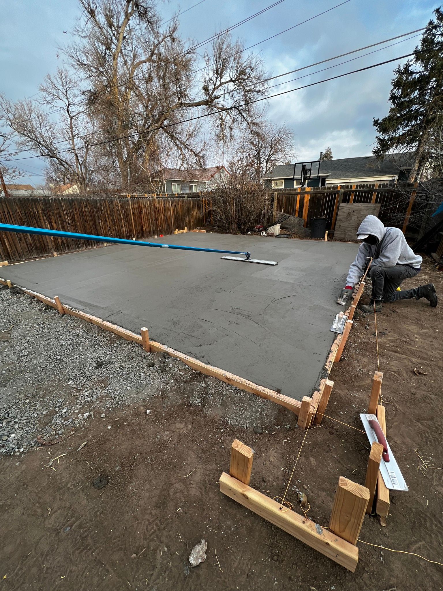 A person in a hooded sweatshirt kneels to finish the edge of a freshly poured concrete slab within wooden form boards.