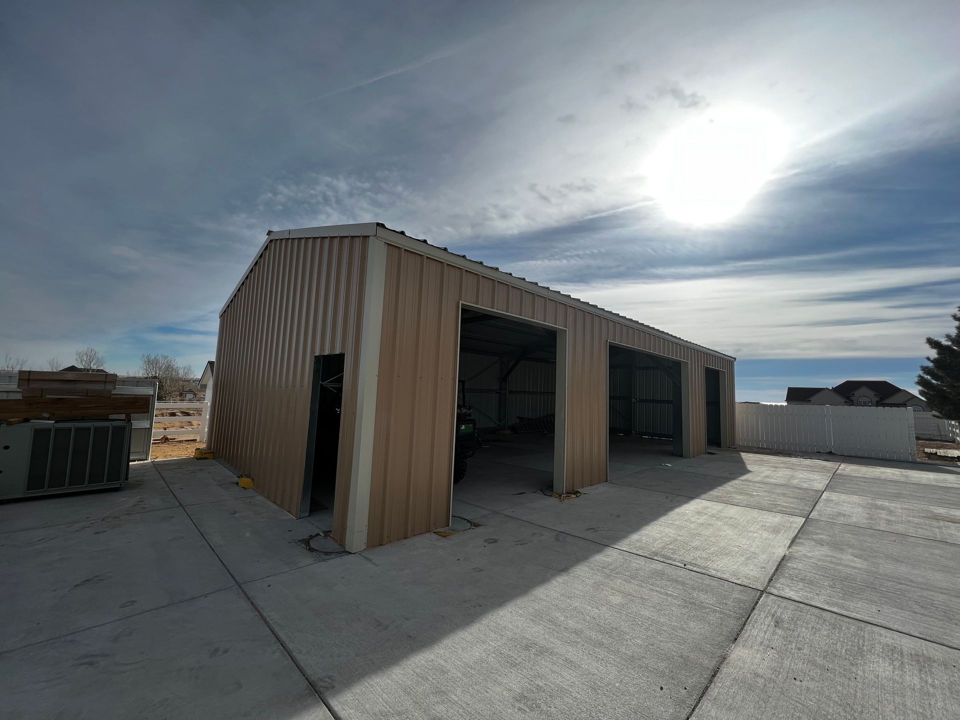 A tan metal garage with two open vehicle bays and a side door sits on a concrete pad under a bright, sunny sky.