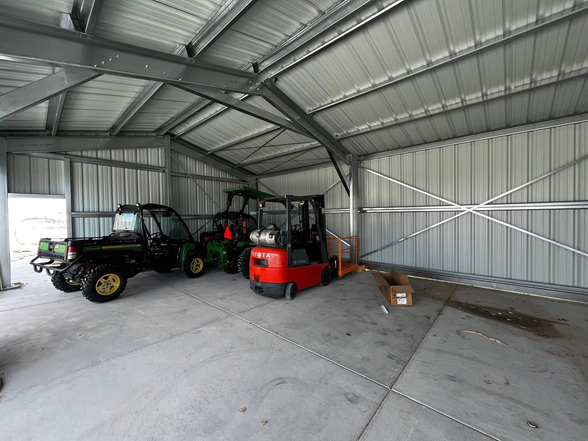 A large metal storage shed containing a green utility vehicle, a red forklift, and a small cardboard box on the floor.