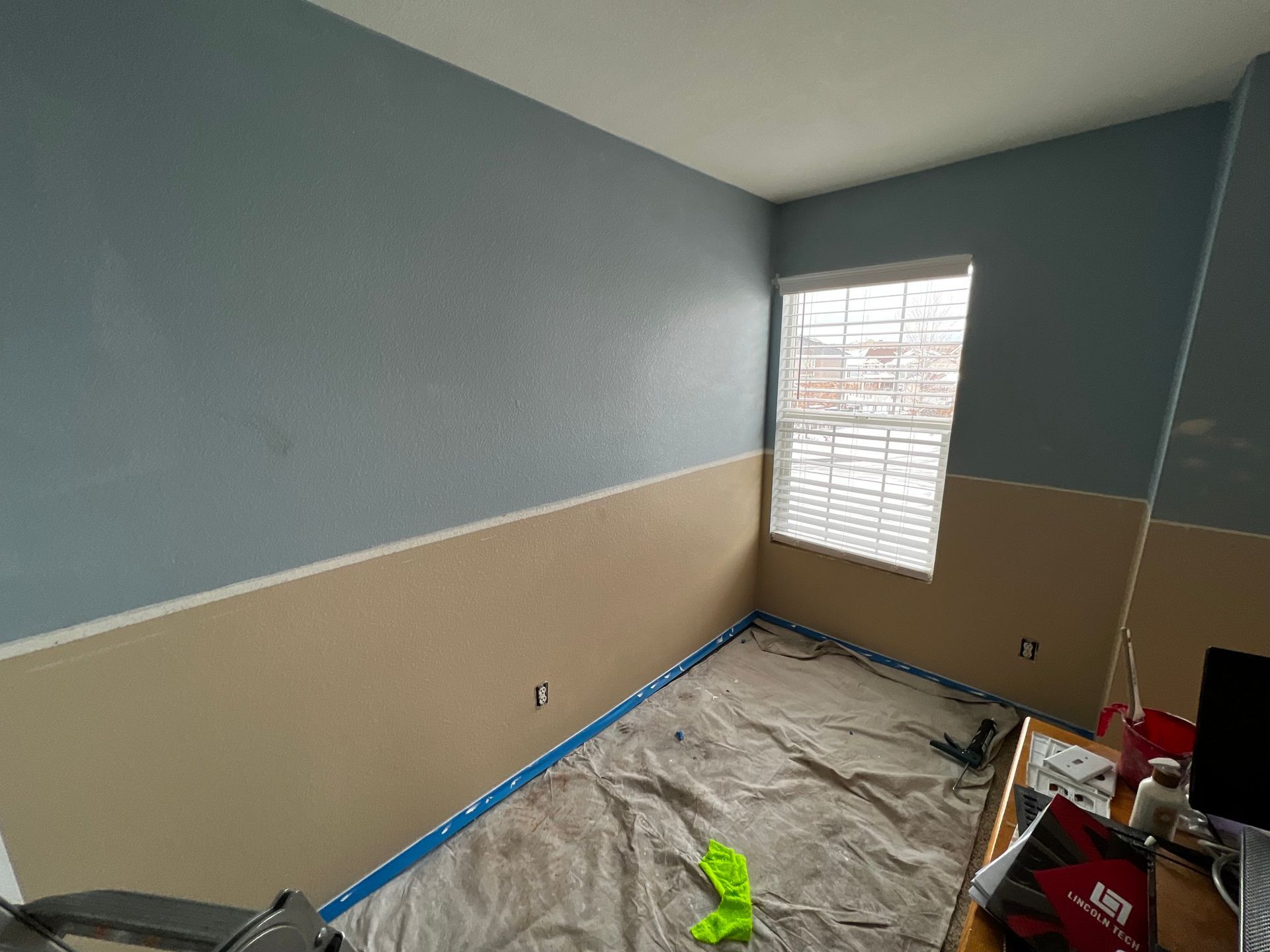 A room under renovation with blue and beige walls, blue painter's tape, and a drop cloth covering the floor.