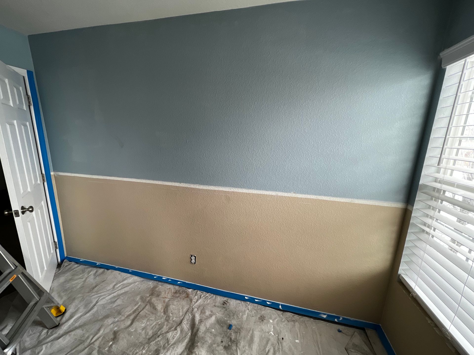 A room wall undergoing a two-tone paint job, with light blue on top, tan on bottom, and painter's tape along the edges.