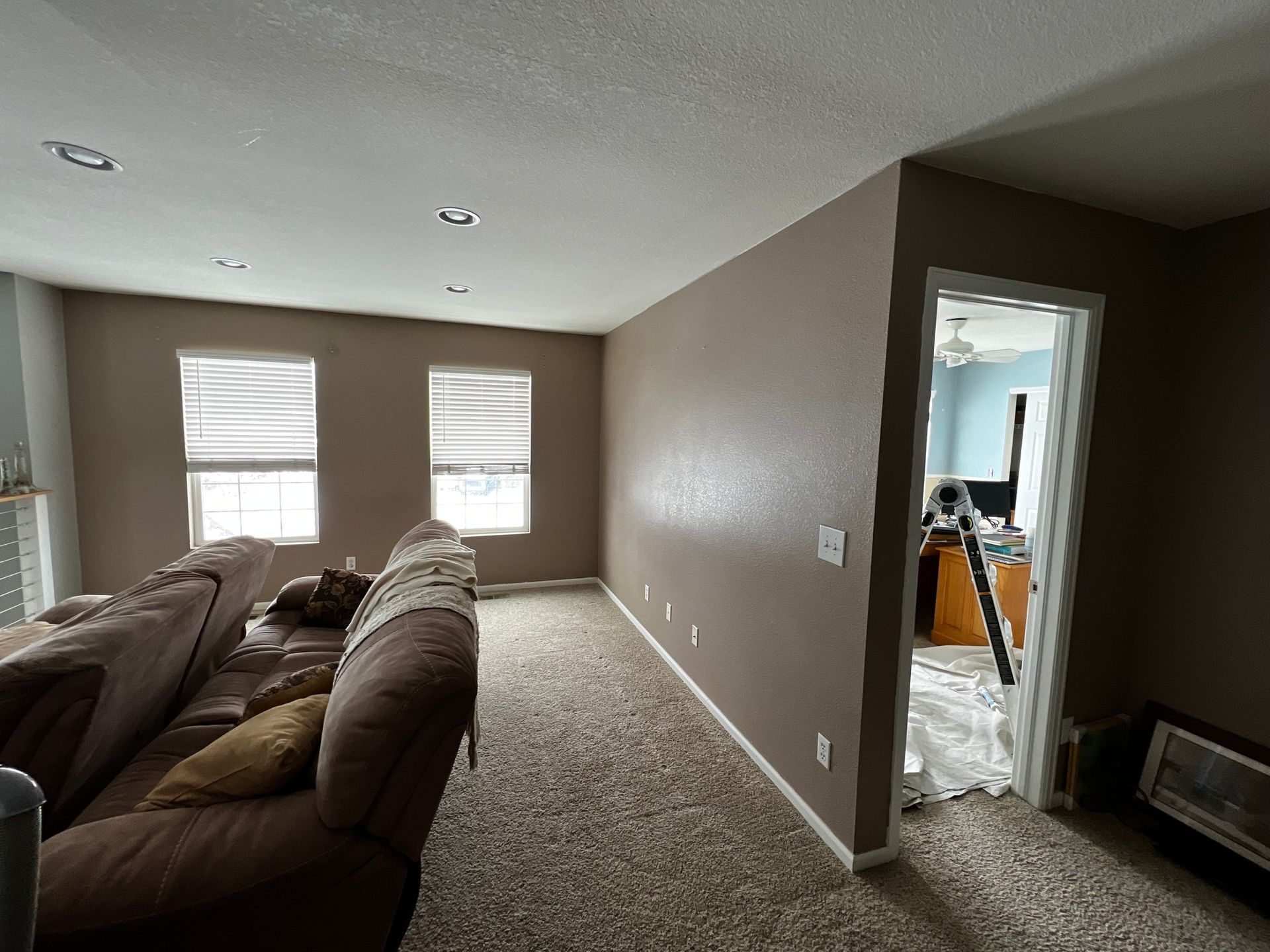 A brown carpeted room with a sofa facing two windows, and an open doorway leading to a light blue room with a desk.