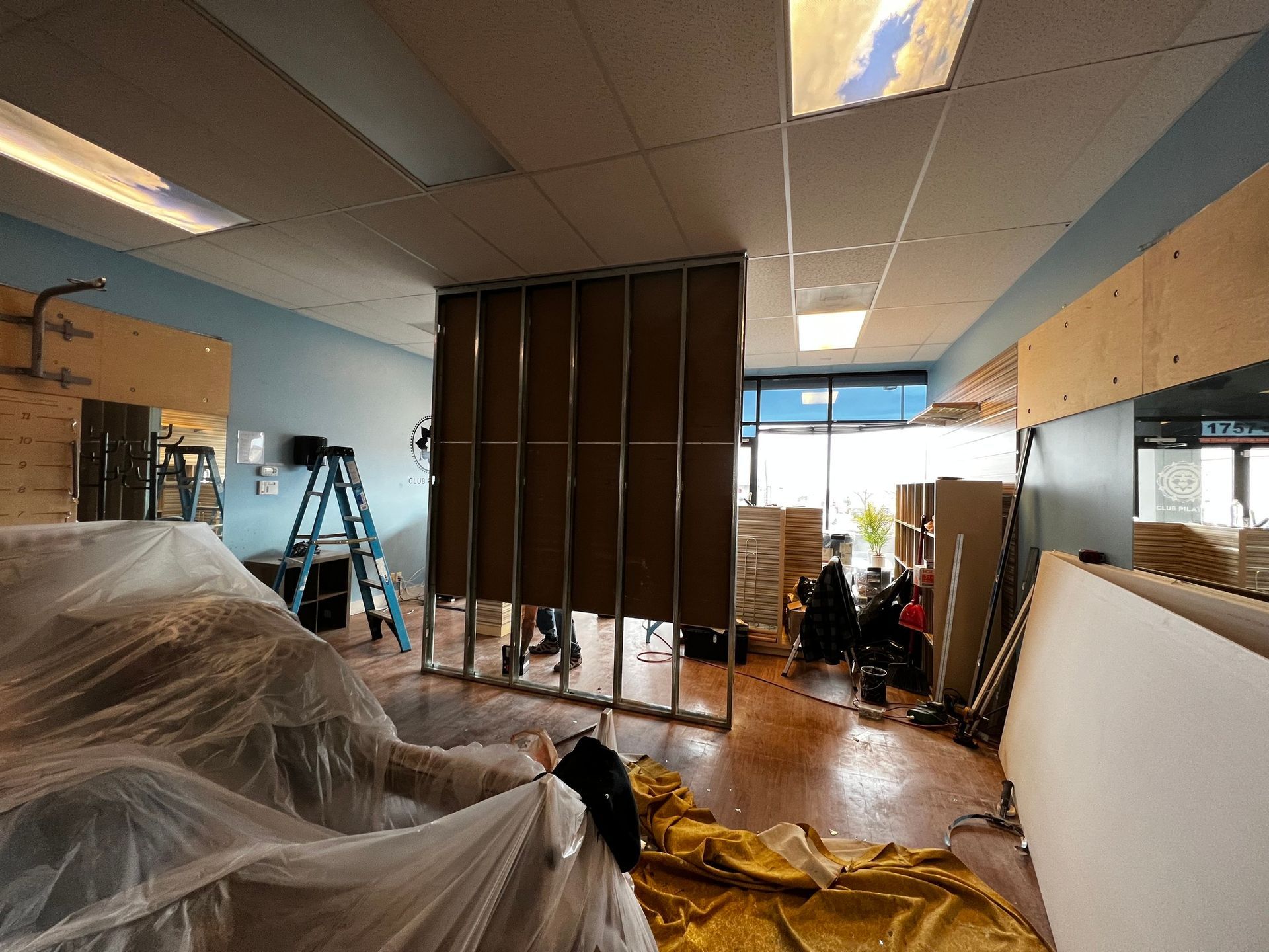 An office space under construction with a metal stud wall, a step ladder, drop cloths, and loose plywood sheets.