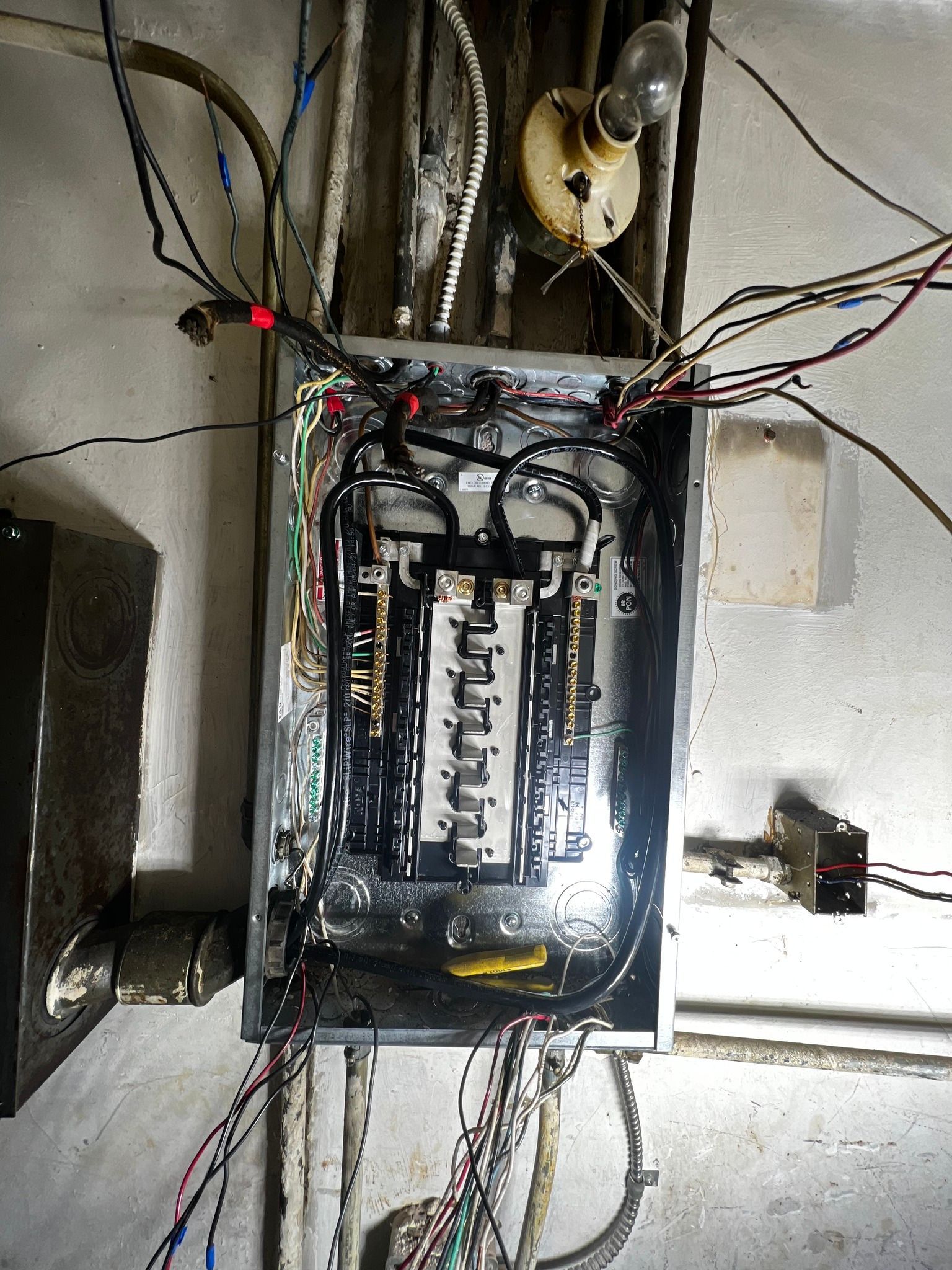 An open electrical breaker panel with exposed wiring and a light bulb fixture mounted on the wall above it.