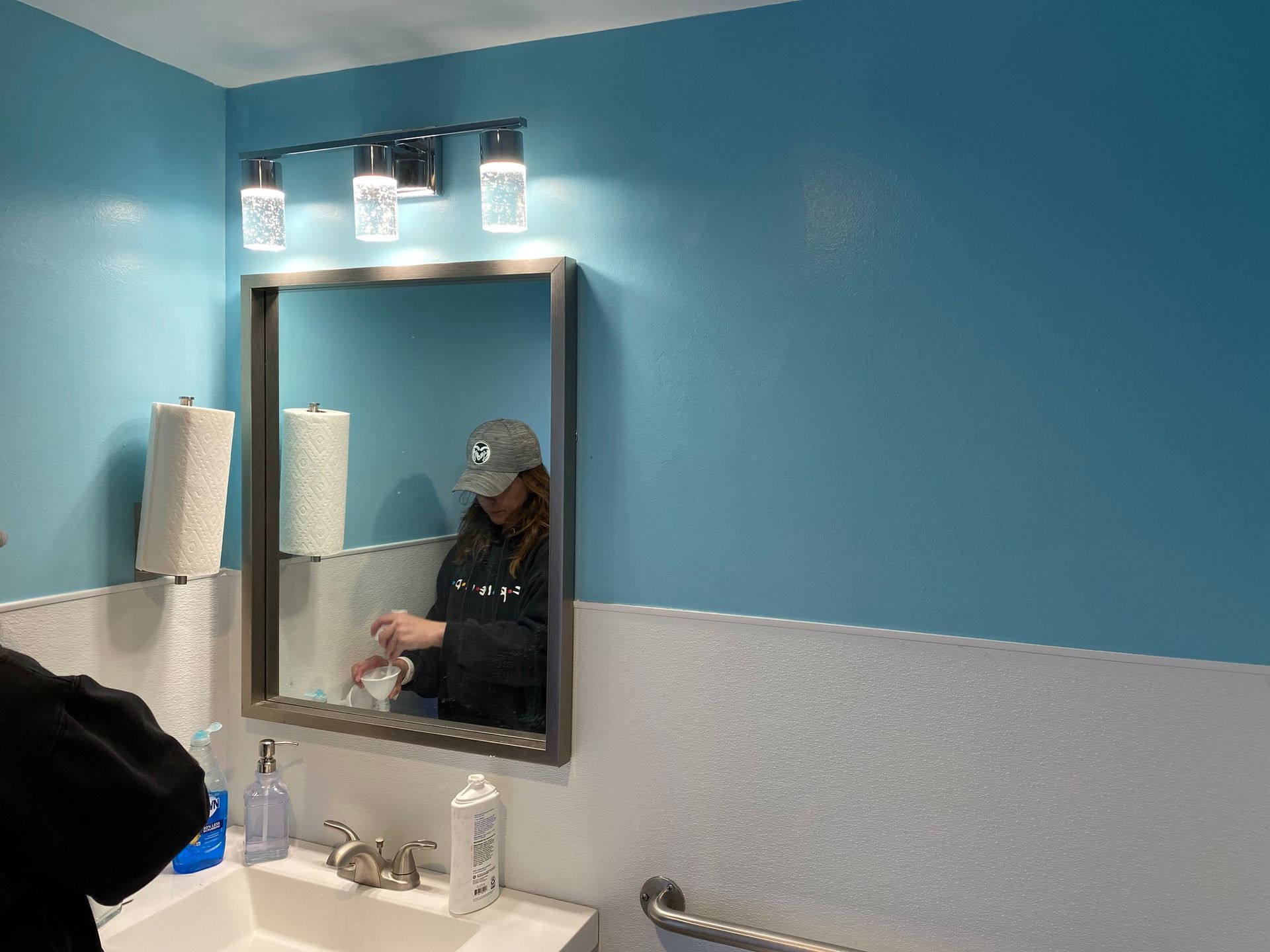 A bathroom mirror reflects a person wearing a cap, above a sink with soap dispensers and a roll of paper towels.