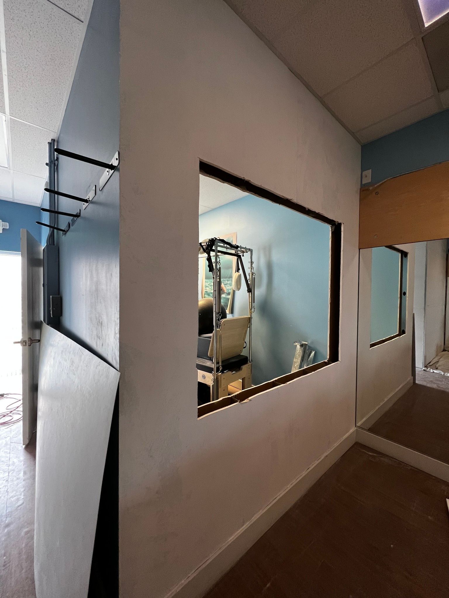 A white wall with a square window looking into a room with a medical lift, adjacent to a large wall mirror.