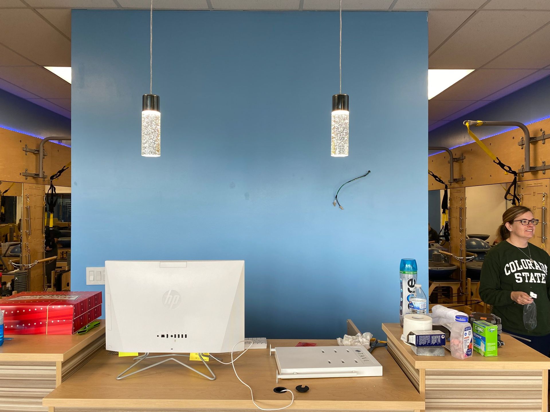A reception desk with a computer faces a blue accent wall with two pendant lights, as a person stands nearby in a gym.