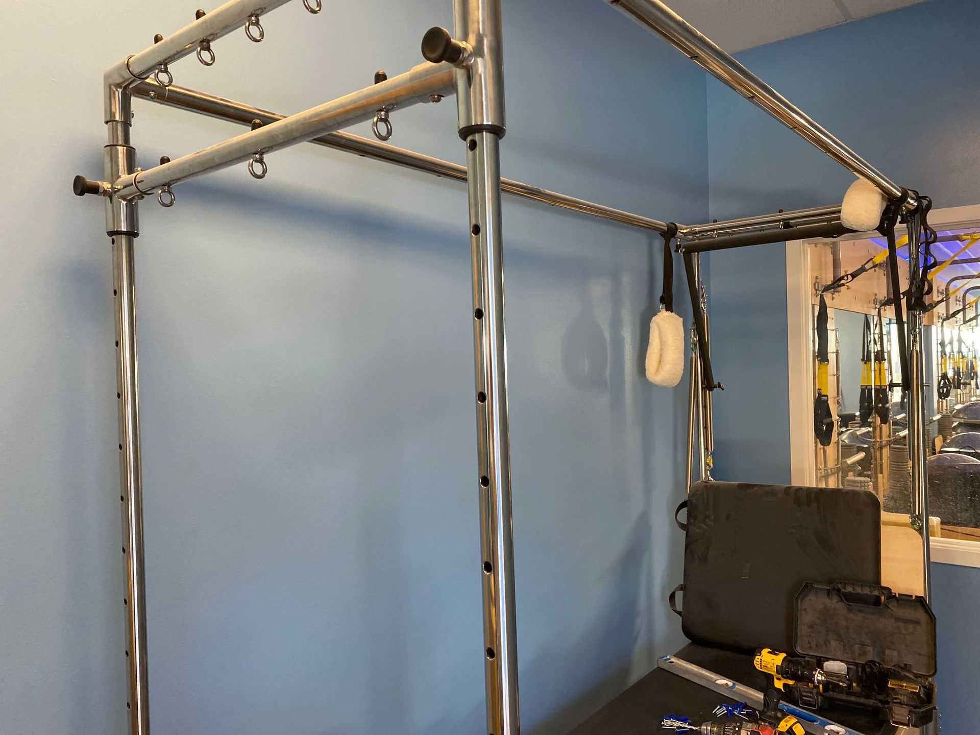 A metal Pilates Cadillac exercise table with hanging straps and a black mat against a light blue wall.